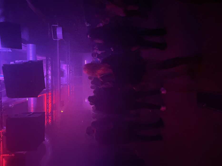 The image depicts an indoor scene within a crowded venue, identified as being in Paris, France. The environment is heavily illuminated by artificial red and purple lighting, with a visible atmospheric haze or smoke effect. The ceiling features exposed industrial elements including large, rectangular black sound speakers suspended from above, horizontal metal truss structures emitting red light, and visible ventilation ducts.
Numerous individuals are present, filling the midground and background of the scene. Most people are rendered as dark silhouettes due to the low light and backlighting conditions, making individual features and precise actions indistinguishable. They appear to be standing or moving within the space, dressed in dark attire. In the right-center foreground, a person with reddish-orange hair is seen from behind, wearing dark clothing. This individual exhibits slight motion blur. The floor is dark and unremarkable.