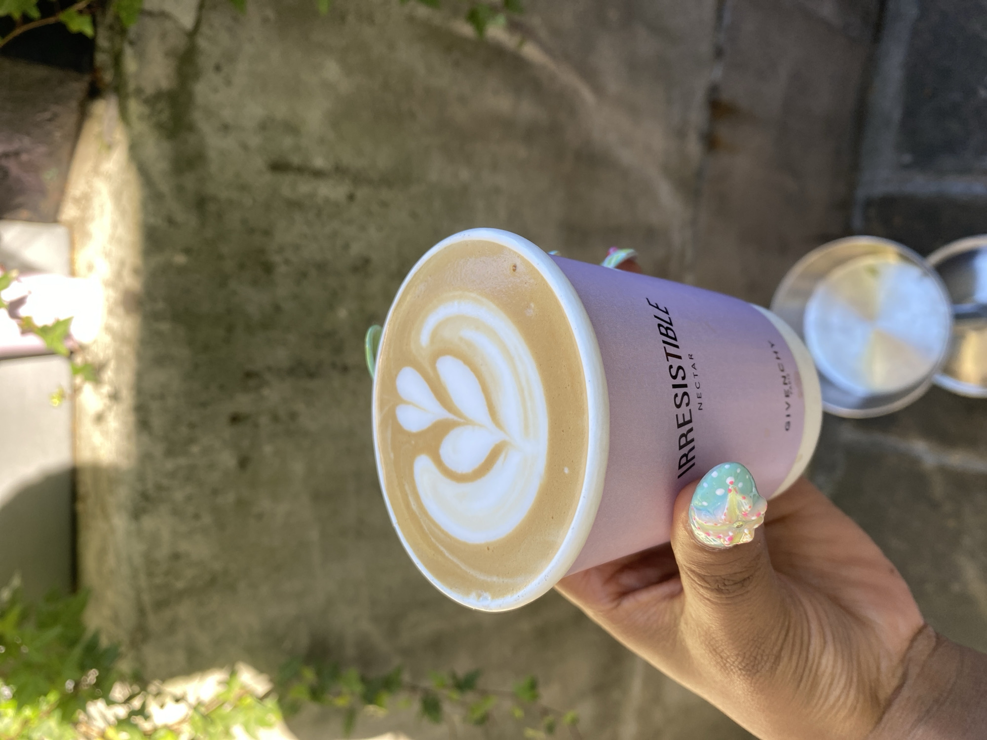 A close-up shot captures a person's hand holding a lavender-colored coffee cup horizontally in an outdoor setting in London, United Kingdom. The hand, with a medium skin tone, features a decorated thumbnail with light blue or mint green polish, white polka dots, and pink and yellow glitter.
The paper coffee cup displays "IRRESISTIBLE NECTAR" and "GIVENCHY" printed vertically in black text on its side. The surface of the coffee within the cup showcases well-executed latte art, forming a delicate pattern of interconnected white hearts or leaves against the caramel-brown beverage.
The background is softly blurred, revealing a textured wall, possibly stone or concrete, partially covered with green ivy or other foliage. Bright natural light illuminates the scene, suggesting it is daytime and possibly sunny. Metallic objects, possibly lids or coffee preparation tools, are faintly visible in the blurred background to the right. The image conveys a pleasant, leisurely moment enjoying a crafted coffee outdoors.