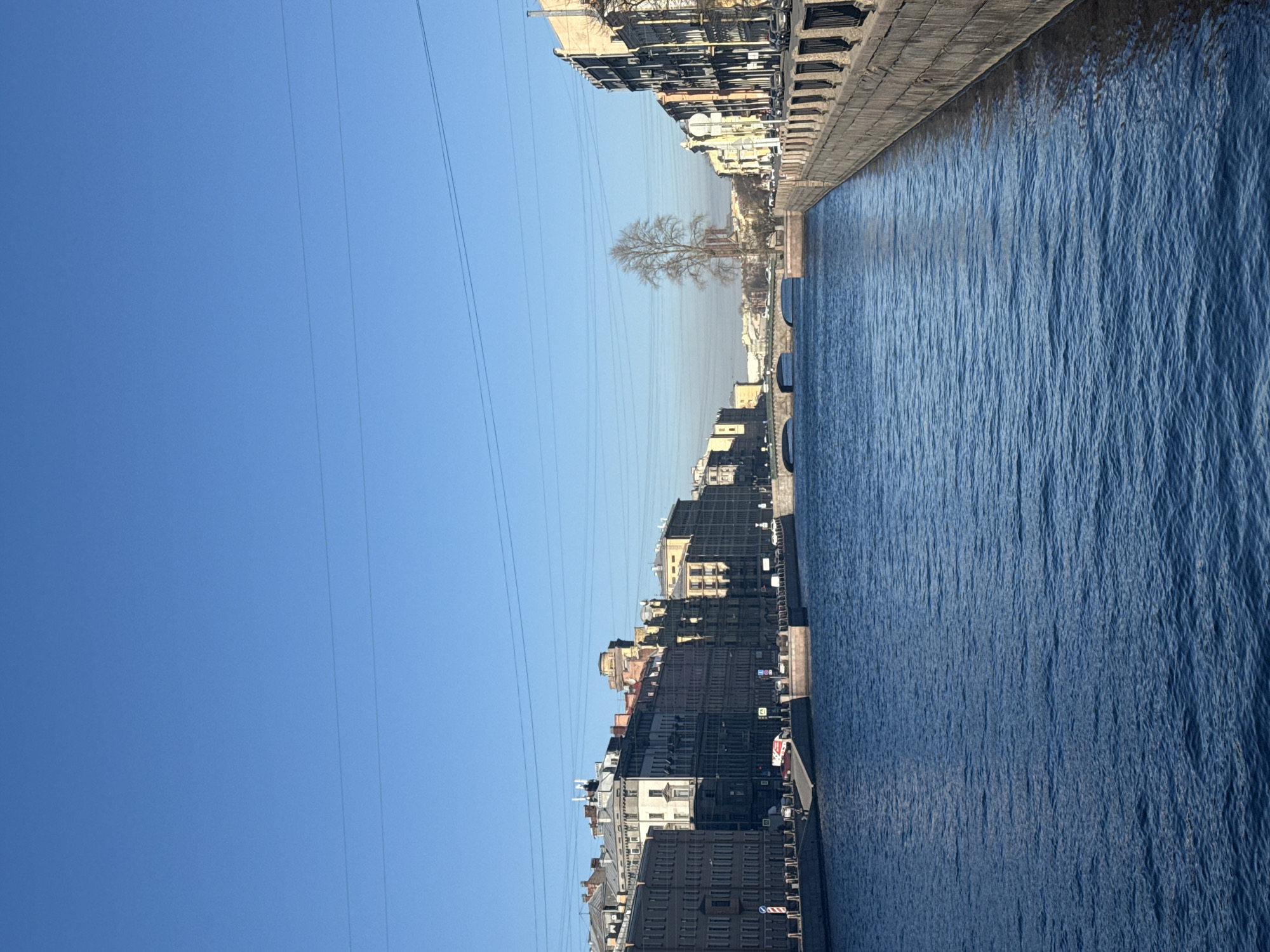 The image shows a view along a canal in Saint Petersburg, Russia. On either side of the canal are buildings, mostly residential and commercial, with varying architectural styles. The canal water is a dark blue with ripples and reflections of the buildings and sky. Overhead power lines stretch across the sky above the buildings. The sky is a clear, bright blue. A bare tree stands in the distance between buildings. The photo is taken during the day with sunlight illuminating the scene.