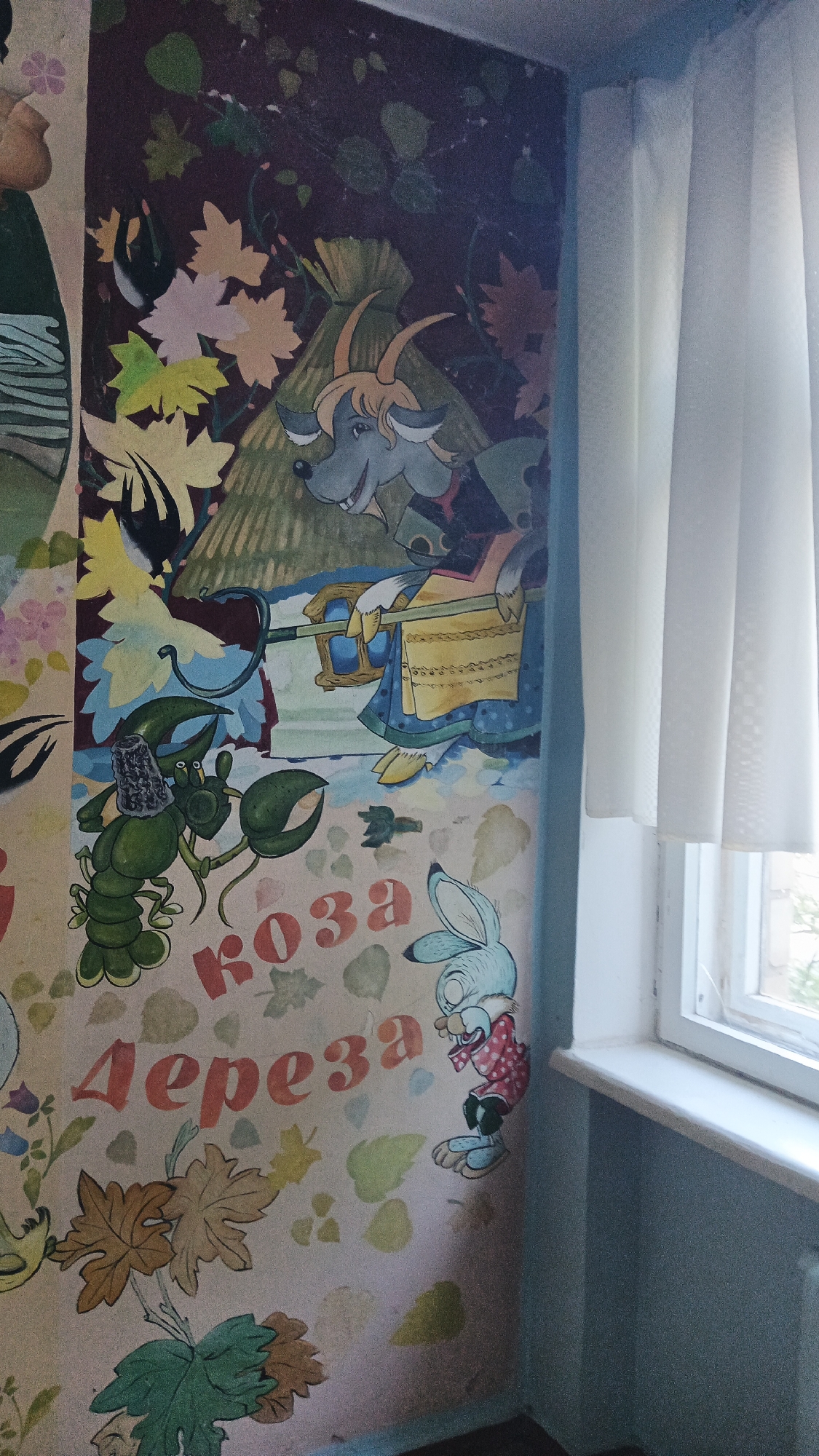 The image shows a mural painted on a wall, likely in a public space such as a school or community center, in Kryvyi Rih, Ukraine. The mural features cartoon-like depictions of animals and plants. In the upper portion of the mural, a goat-like creature with horns and a blue face is depicted. To its left, a green lobster-like creature wearing a hat is shown. Below these characters, the Russian words "КОЗА ДЕРЕЗА" are painted in red lettering, which translates to "Goat Dereza," a character from a Russian folk tale. To the right of the lettering, a blue rabbit character wearing a red polka-dot shirt is depicted. The background of the mural consists of stylized leaves and floral elements. The wall is adjacent to a window with white curtains, and a windowsill is visible. The overall condition of the mural suggests it is an older piece, with some wear and tear visible.
