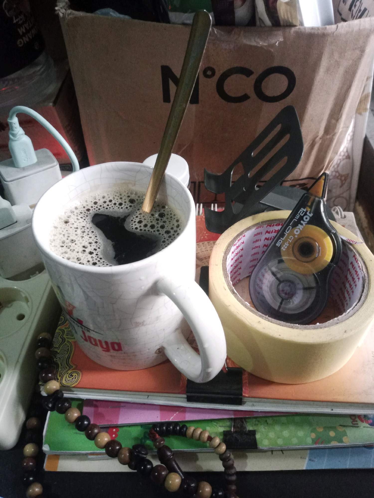 The image depicts a tabletop scene. In the foreground, a white mug filled with dark liquid and topped with foam is situated on top of several books. A wooden stirrer is visible in the mug. To the right of the mug, a roll of yellow tape is present, with a white correction tape dispenser resting on top of it. Behind these objects, a large cardboard box with the "M.CO" logo is partially visible. To the left of the mug, a white electrical adapter with a light blue cord is plugged into a power outlet. A string of dark brown and light brown beads is draped across the lower portion of the image, partially obscuring the books. The scene appears to be indoors, possibly in a home or office environment. There is no indication of the specific location within the city of Balaraja, Indonesia.
