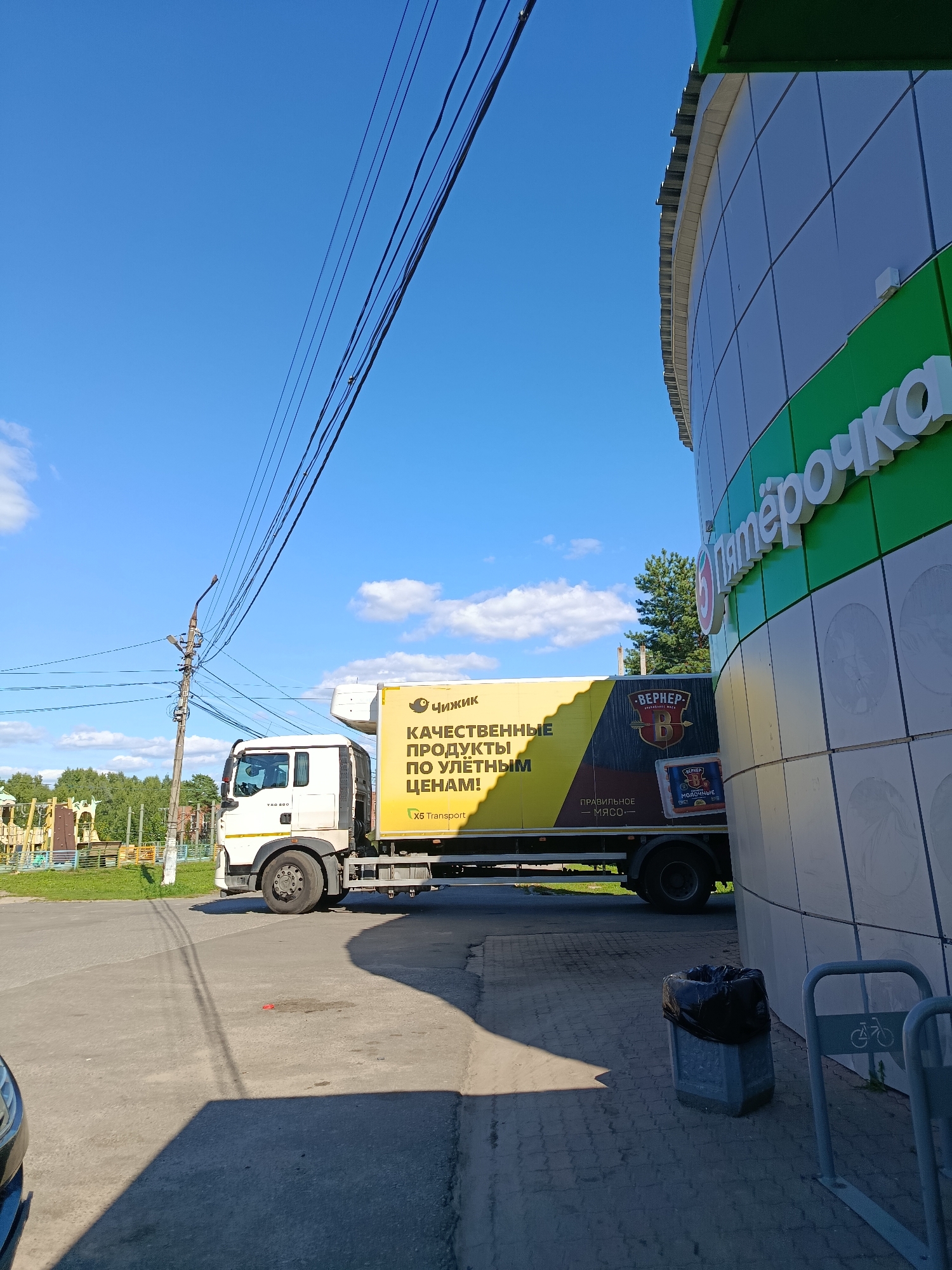 An eye-level, medium shot captures a white truck with a yellow trailer parked on a paved surface outside a modern building with a curved facade in the city of Vereya, Russia. The building has a prominent green and white logo that reads "Пятёрочка." The truck's trailer is emblazoned with text in Russian, including "ЧИЖИК" (Chizhik), "КАЧЕСТВЕННЫЕ ПРОДУКТЫ ПО ДОСТУПНЫМ ЦЕНАМ!" (Quality products at affordable prices!), and "X5 Transport." A logo for "ВЕРНЕР" (Werner) is also visible on the trailer, along with an image of milk products. In the background, a playground with climbing structures and swings is visible, bordered by trees. The sky is bright blue with scattered white clouds. Overhead, a network of dark power lines is strung across the frame. A black trash bag is placed in a concrete bin near the building entrance, and bicycle racks are present.