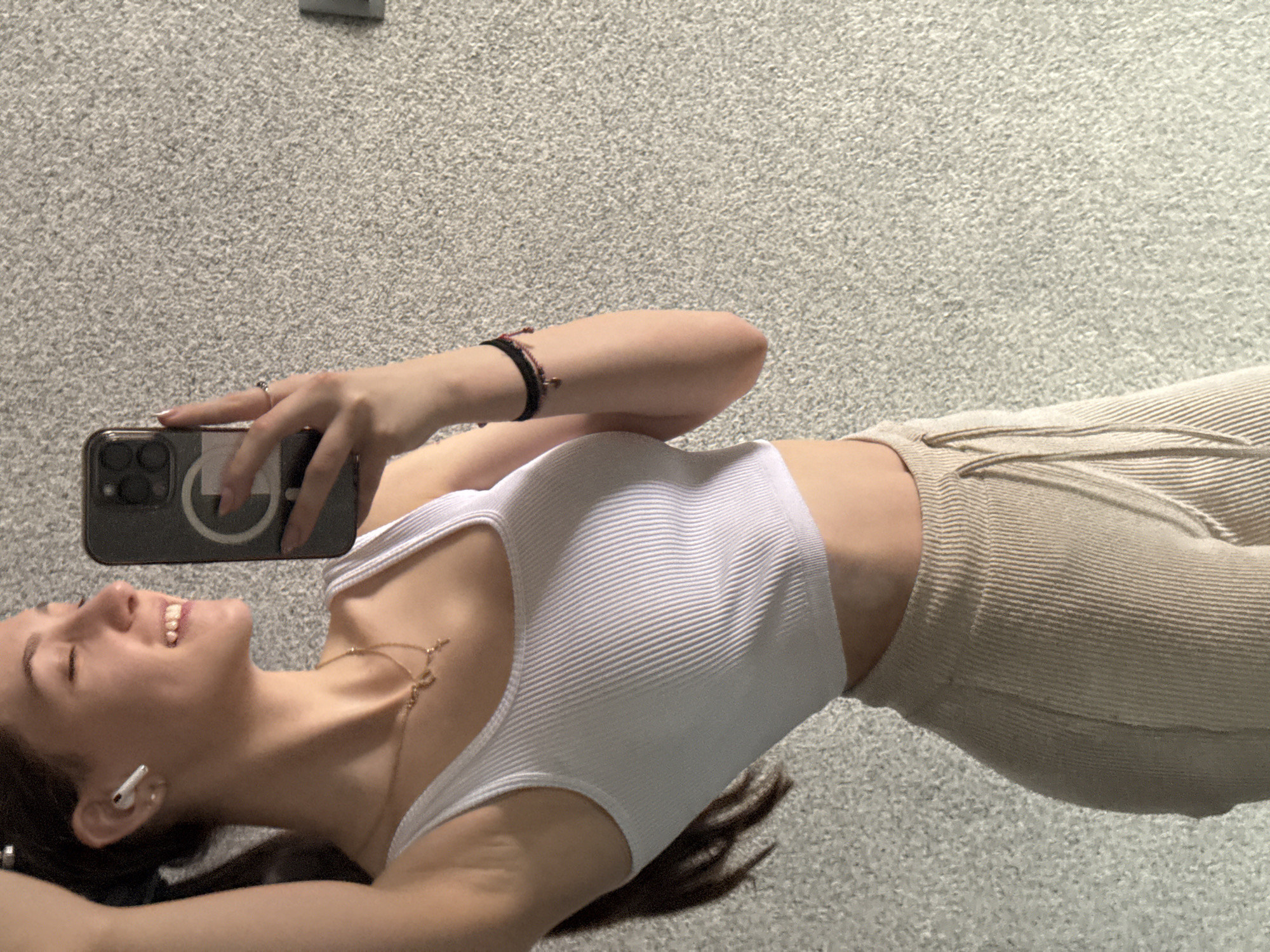 The image displays a young adult female captured in a selfie, taken indoors in Sofia, Bulgaria. She is positioned against a light gray, heavily textured wall. The subject is smiling broadly with her eyes nearly closed, revealing her teeth. Her dark hair is partially visible, with her right arm raised as if adjusting her hair. She is wearing a white ribbed crop top and light beige ribbed pants. Accessories include a white wireless earbud in her left ear, a gold-colored layered necklace with a cross pendant, and a dark-colored bracelet with a charm on her right wrist. A ring with a dark stone is visible on her left ring finger. Her left hand holds a dark-colored smartphone, likely an iPhone, directed towards herself. A white rectangular electrical fixture, possibly a light switch or outlet, is partially visible on the wall to the right of the frame.