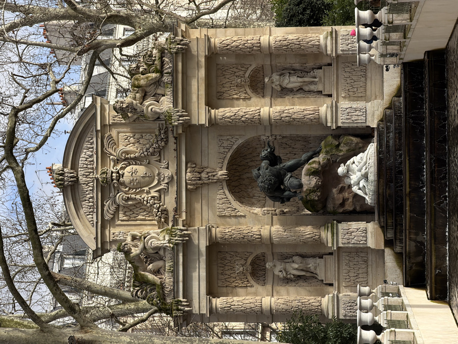 This image captures the Medici Fountain, a prominent stone monument in the Jardin du Luxembourg, Paris, France, under a clear daytime sky.
The central feature is a large, ornate, multi-tiered classical fountain structure constructed from light-colored stone, exhibiting areas of green patina and moss. At its uppermost level, a central escutcheon featuring a crowned fleur-de-lis motif is flanked by two reclining classical figures. Below this, the main section of the fountain features three arched niches separated by fluted columns. The central and largest niche houses a dark bronze sculpture depicting Polyphemus, a cyclops, standing aggressively on a rock, observing the white marble figures of Galatea and Acis embracing on a lower rock formation. Flanking this central tableau, the two smaller side niches each contain a standing white marble classical statue, one male on the left and one female on the right.
Water flows from the central sculpture, cascading down several dark-colored, shallow steps into a lower basin, creating ripples. A stone balustrade with decorative white urns lines the sides of the water feature.
The fountain is partially framed by the bare, intricate branches of large deciduous trees, suggesting winter or early spring. Beyond the trees and above the fountain, typical Parisian buildings with light facades, dark rooftops, and chimneys are visible in the background. On the far right, partially obscured by the balustrade, one individual wearing a bright pink or red hat is visible. The scene is illuminated by natural light, casting distinct shadows.