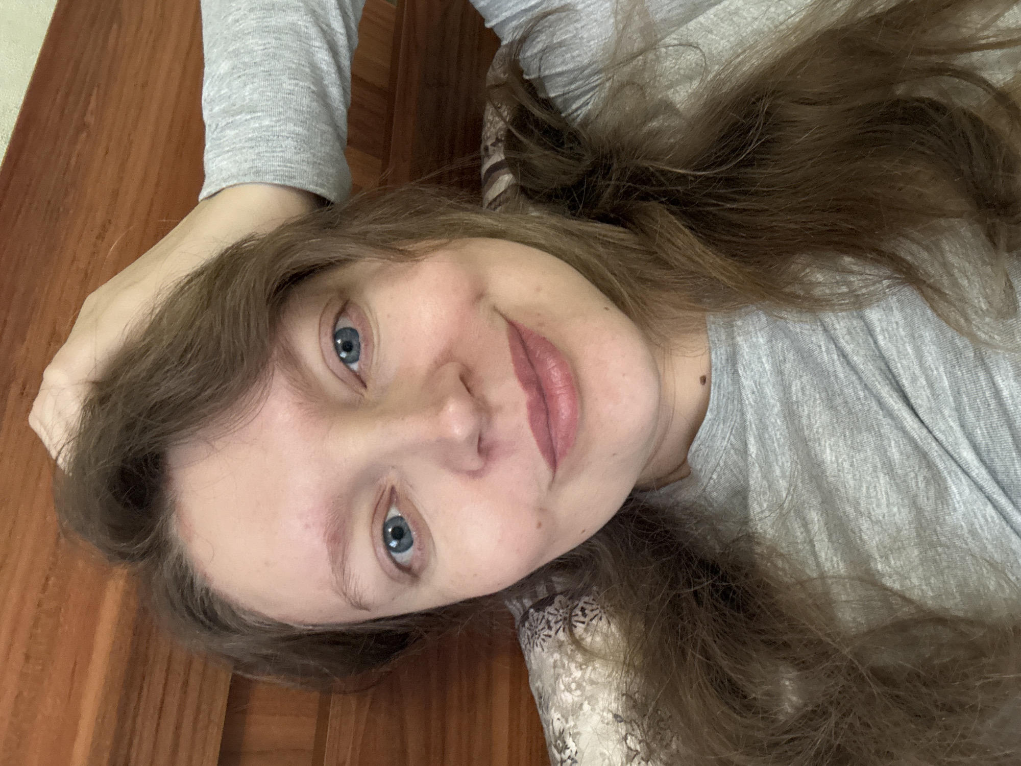 A fair-skinned woman with long, light brown hair and blue eyes is lying down, facing the camera. She is wearing a gray long-sleeved shirt. Her head rests on a wooden surface. A patterned material is visible beneath her head, possibly a pillow or cushion. The background is a plain wooden surface. There is no information to determine if the photo was taken in Sludka, Russia.