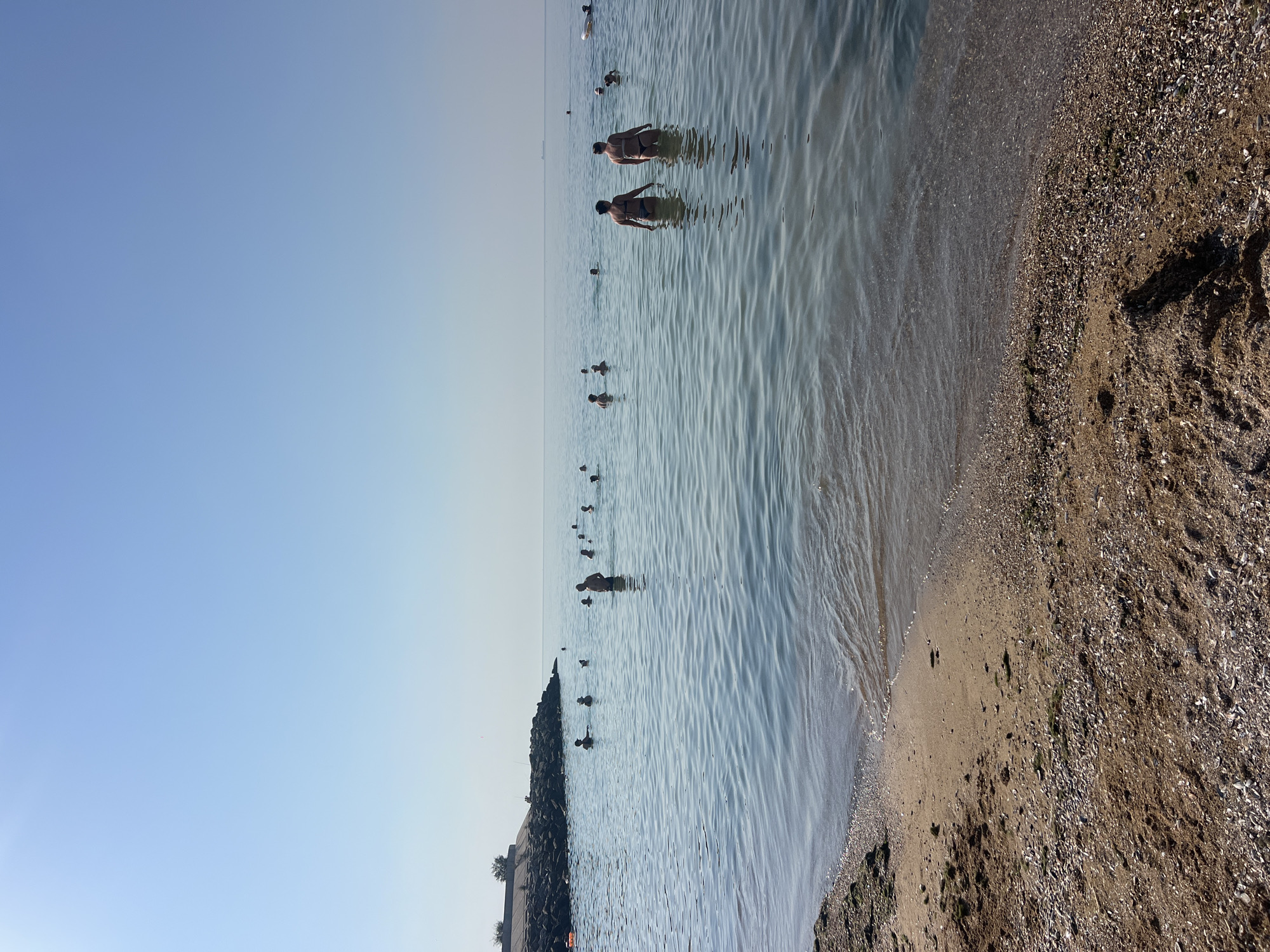 The image shows a group of people bathing in the sea on a sunny day, with a clear blue sky overhead. The scene appears to be a beach on the Black Sea, likely in the Fontanka, Ukraine area, given the presence of a man-made breakwater or pier structure on the left side of the image, typical of coastal defenses or access points in the region. The water is calm and shallow near the shore, with small waves lapping onto a sandy beach covered with shells. Several individuals are visible in the water, some wading and others swimming. In the distance, a ship can be seen on the horizon. The overall atmosphere is one of leisure and relaxation.
