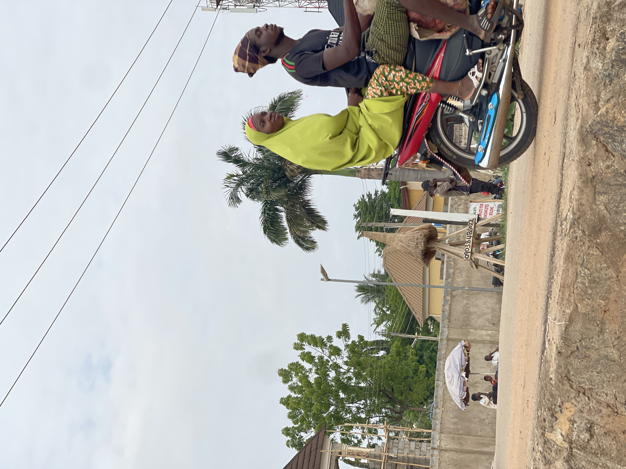 An outdoor scene in Abuja, Nigeria, depicts a busy street on an overcast day with soft, diffused lighting. The main subjects are three people riding a red and silver motorcycle. The driver, a man, wears a dark t-shirt with a white "Emirates" logo and a patterned headwrap, looking upwards. In front of him sits a woman dressed in a vibrant lime green hijab and matching garment, looking forward. A third passenger, wearing green and white checkered fabric, is partially visible behind the driver.
The motorcycle is positioned on what appears to be a paved road, adjacent to a dirt or gravel shoulder that occupies the right foreground. Overhead, multiple power lines stretch across the pale, cloudy sky. A large palm tree stands prominently behind the motorcycle. In the middle ground, various other green trees, buildings with reddish-brown roofs, and structures are visible. One notable structure features a thatched-like roof supported by poles. A partially constructed building with concrete blocks and wooden scaffolding can also be seen.
Further in the background, several individuals populate the scene. One man stands near the rear wheel of the motorcycle, wearing a dark cap. To the right, three people are gathered under a white umbrella or canopy, suggesting a small market stall or resting area. A partially legible white sign in the background displays text including "TO LET" and a phone number starting with "0903". The overall atmosphere is calm, capturing a typical moment of daily life and transportation in the area.
