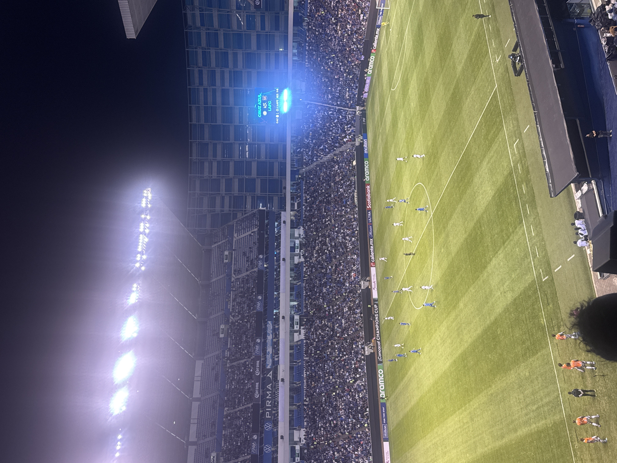 The image depicts a nighttime soccer game at a crowded stadium. The stadium is in Puebla, Mexico, based on the team names displayed on the scoreboard. The score is 1-0 in favor of Cruz Azul against LAFC. A lit stadium scoreboard is visible. The soccer field is illuminated by stadium lights. The stands are filled with spectators. The game is in the 63rd minute. Players are visible on the field. Various sponsor banners are visible along the side of the field.