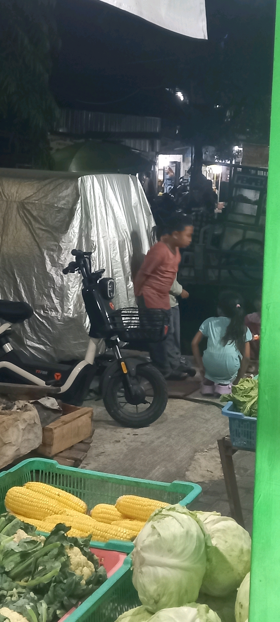 This is a vertical, medium-distance, full-body shot of a boy standing next to an electric scooter in Sedati Agung, Indonesia. The boy, wearing a red t-shirt and dark pants, is looking to his right, away from the camera. He stands to the right of a black and white electric scooter, which has a basket on the front. Behind the scooter, a large tarp is draped, obscuring much of the background. To the right of the boy, a young girl wearing a light blue dress and pink shoes is squatting, also looking to her right. In the foreground, to the left, a green plastic basket contains several ears of corn and other vegetables. To the right, another green basket holds several heads of lettuce. The scene appears to be outdoors, possibly a market stall, with a concrete ground surface visible. The lighting suggests it is evening or nighttime.