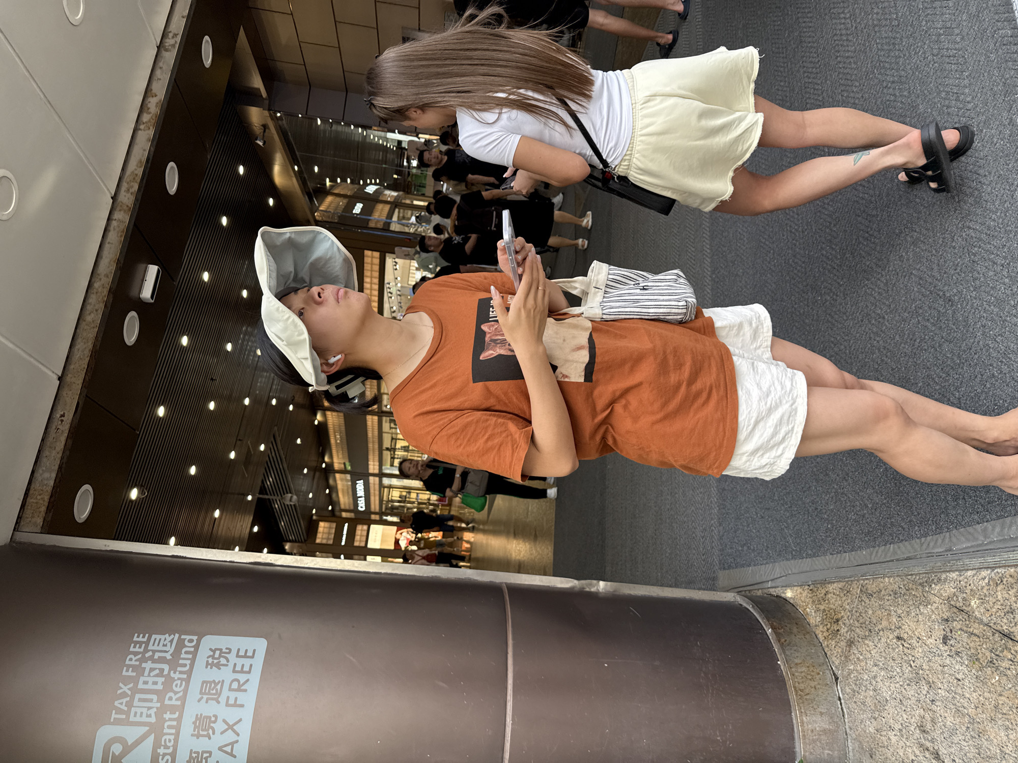 A medium shot captures two young women standing outside a building in Haizhu District, China. The woman on the left, facing mostly away from the camera, wears an orange t-shirt with a graphic print of a cat, white shorts, and a wide-brimmed white sun visor. She has earbuds in and is holding a smartphone. A canvas tote bag with vertical stripes hangs from her shoulder. The woman on the right is seen from the back, wearing a white t-shirt, light yellow shorts, and black platform sandals. She has long, light brown hair and a tattoo of a leaf on her left calf. She is also holding a smartphone. In the background, other people are visible walking past shops with illuminated signs, including "CASA MODA." The environment appears to be a covered walkway or shopping area. A large brown cylindrical pillar with a "TAX FREE" sticker is present on the left side of the frame.