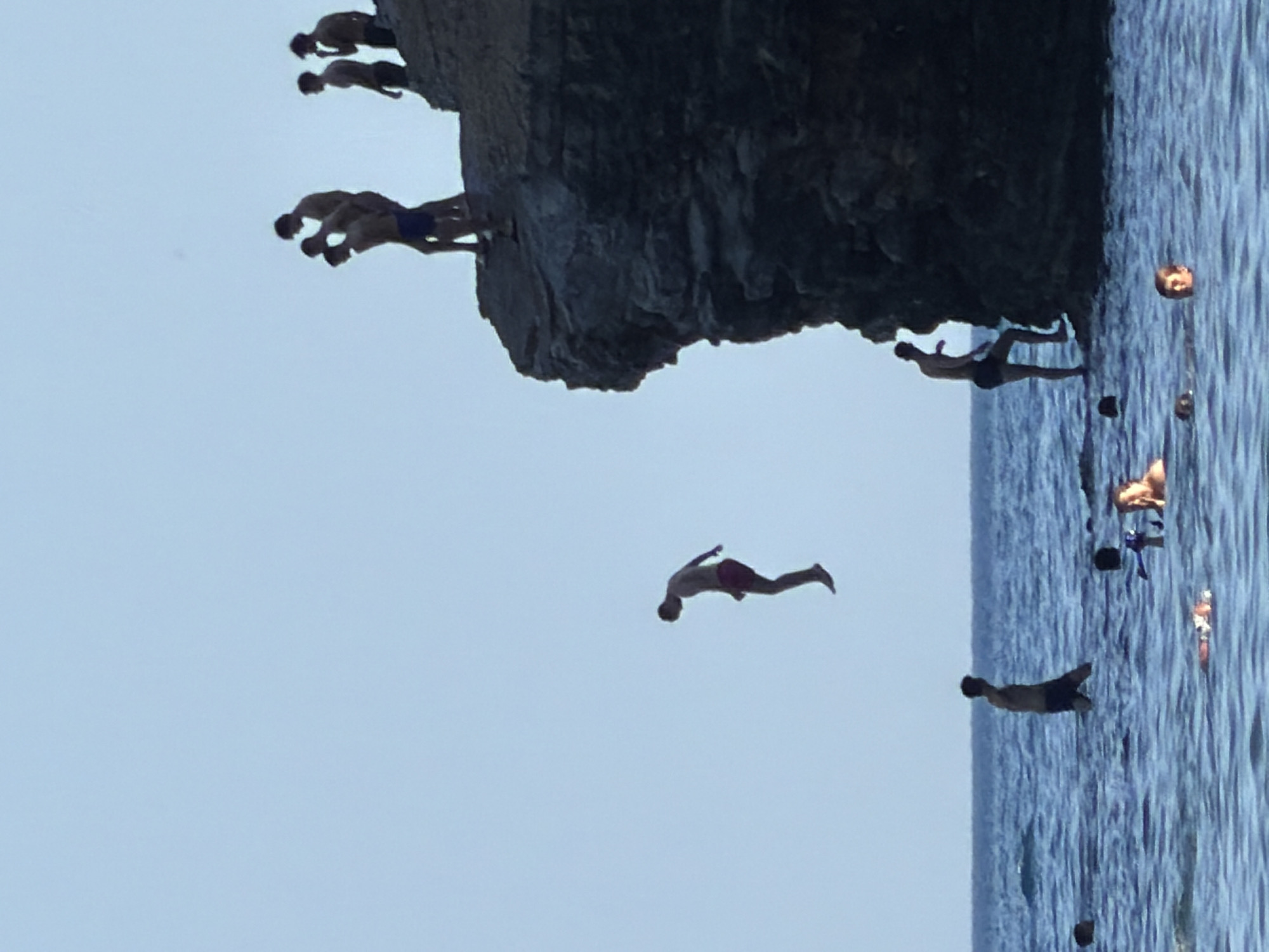 In Budva, Montenegro, a group of young men are engaged in cliff diving from a rocky outcrop into the clear blue sea. The primary focus is on a man in mid-air, captured in the act of diving, with his body angled downwards. Several other individuals are positioned on the edge of the cliff, some appearing to be preparing to jump or observing the divers. In the water below, other people are swimming and enjoying the ocean. The environment is characterized by the imposing cliff face, the expansive sea, and the clear sky.