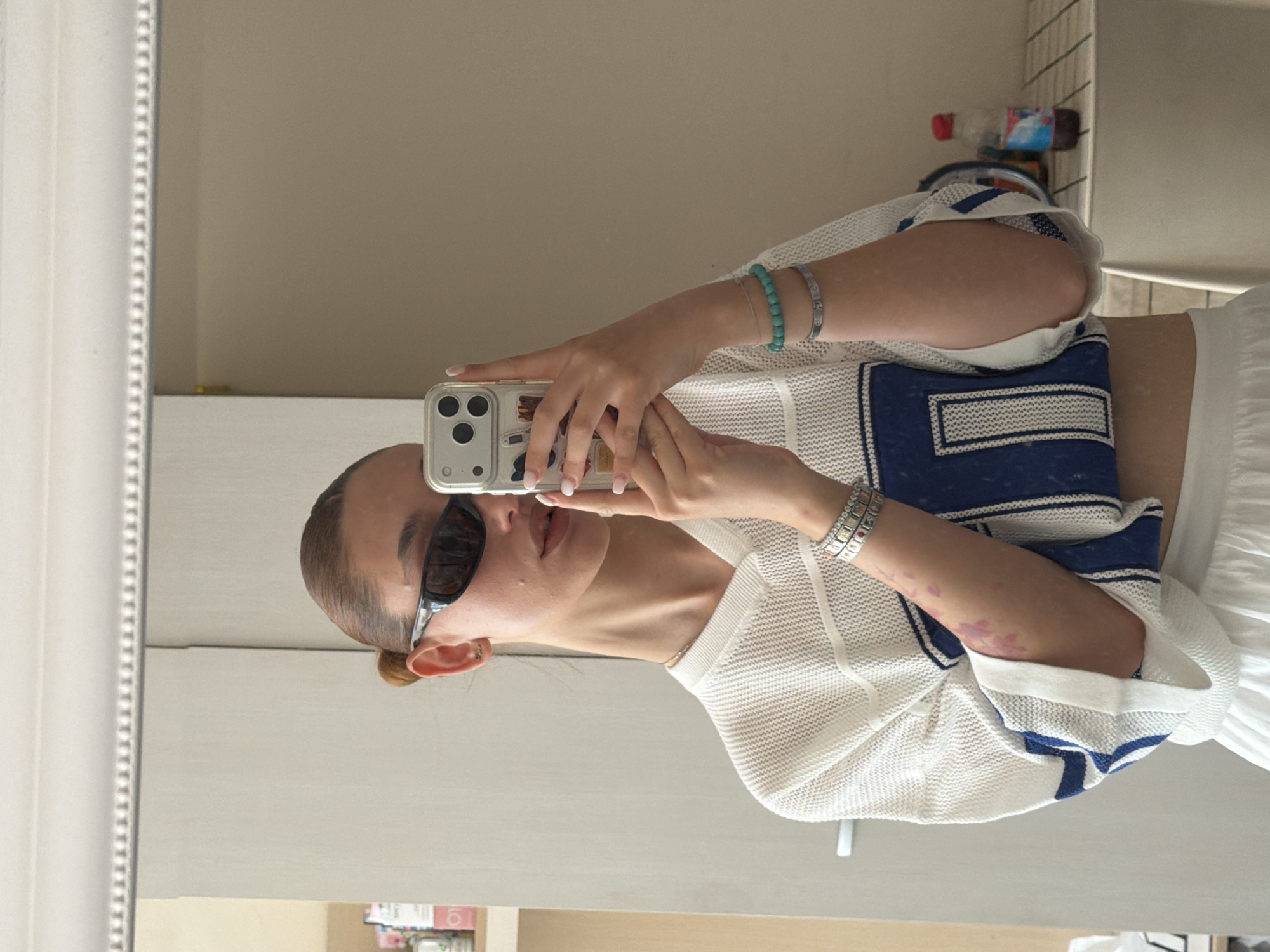 A person is taking a selfie in front of a mirror. The person is wearing sunglasses, a white top with blue trim, and bracelets. The person is holding a smartphone with a clear case and decorative stickers. The background includes a wall, part of a door, and some items on a shelf. It is not possible to determine the location in the city of Sofia, Bulgaria from the image.