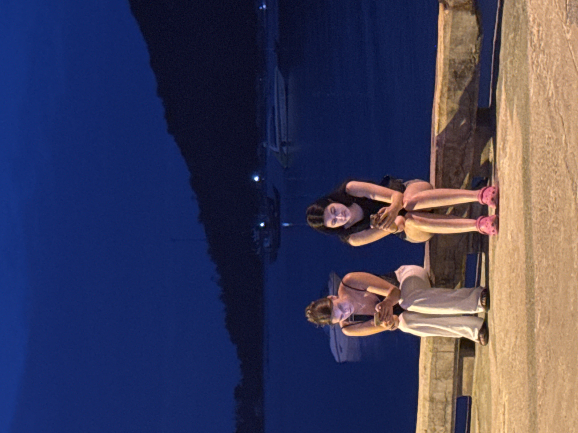 Two young women are sitting on a concrete bench at night, facing forward and looking down at their mobile phones, illuminated by the screens. The woman on the left is wearing a dark tank top and loose white pants, with her hair pulled back. The woman on the right is wearing a dark tank top and shorts, with her long dark hair down. Both women are wearing sandals. They are positioned on a paved surface, with a body of water behind them, which appears to be the sea. In the water, several boats, including a larger white boat with a dark stripe, are visible. The background features a dark, tree-covered hill. The sky is a deep dark blue, suggesting nighttime. The lighting is dim, with the primary illumination coming from the phone screens and ambient light reflecting off the water. The scene appears to be a waterfront area in a tropical location, possibly a pier or promenade, consistent with the coastal environment of Koh Phangan.