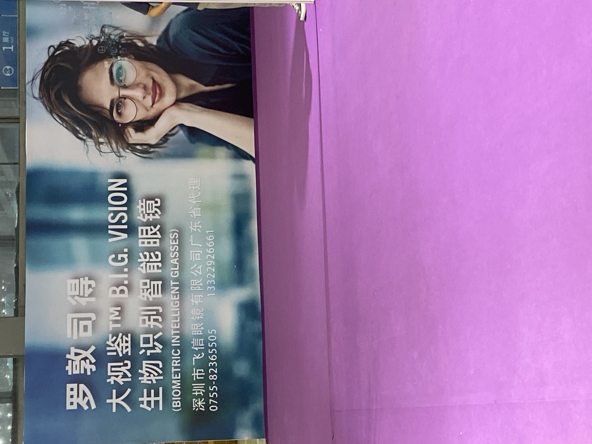 This is a photograph of a large advertisement banner displayed in a public area. The banner features a portrait of a woman wearing eyeglasses, with digital elements superimposed over them, suggesting advanced technology. The text on the banner is in Chinese and English, promoting "B.I.G. VISION" and "BIOMETRIC INTELLIGENT GLASSES." Below the main text, there is information about a company, "Shenzhen Feixin Glasses Co., Ltd. Guangdong General Agent," along with a phone number "0755-82365505" and another number "13322926661." The bottom portion of the image shows a purple floor, likely carpeting, and a shadow cast by the banner. The background behind the woman appears to be a blurred blue and white gradient. The advertisement is likely located in a shopping mall, exhibition hall, or other commercial space within Futian District, Shenzhen, China, given the presence of Chinese text and the company's location mentioned.