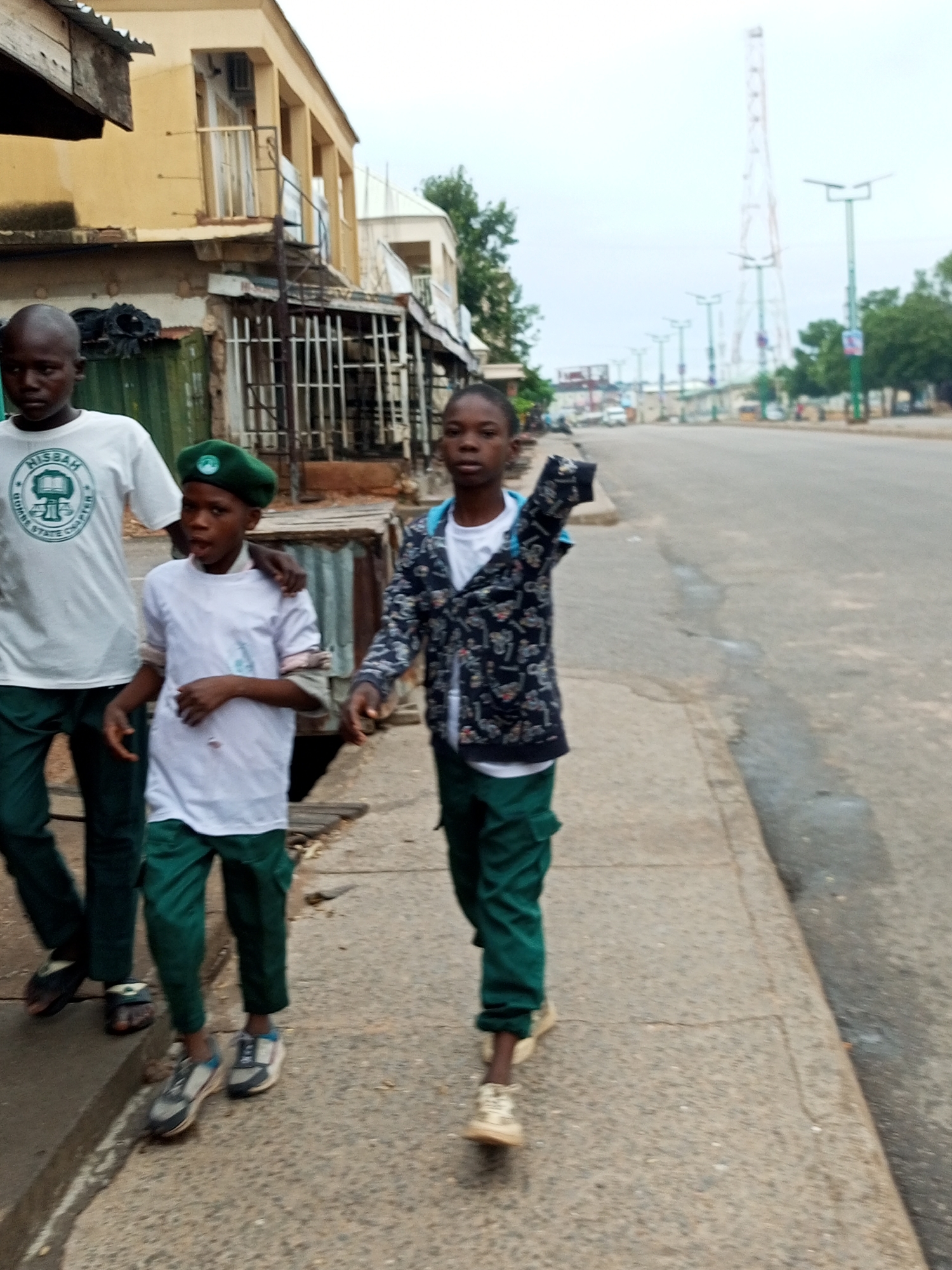 Three young boys walk along a paved sidewalk in Arawa, Nigeria. The boy on the left wears a white t-shirt with a green logo and dark green pants. He is out of focus but appears to be looking forward. The middle boy, wearing a green beret, a white t-shirt, and dark green pants, looks towards the viewer with his mouth slightly open. His left hand is raised and his right arm is around the shoulders of the boy to his right. The boy on the right wears a white t-shirt and dark green pants, and a patterned jacket over his t-shirt. He is also out of focus and walking. The sidewalk is cracked and weathered, with a street to the right. In the background, there are buildings with yellow facades, some with balconies, and a tall communication tower. The sky is overcast.