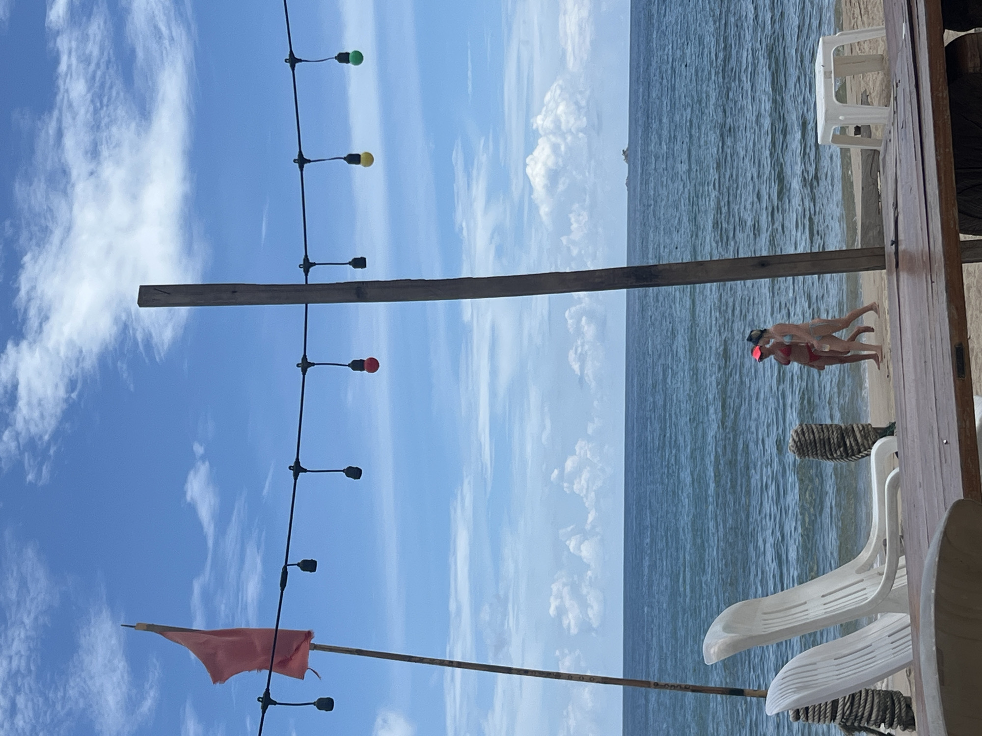 In a beach scene in Choeng Thale, Thailand, two individuals in swimwear walk along the shoreline with the ocean in the background. A string of colorful lights hangs across the frame, with bulbs in red, yellow, and green visible. A red flag is attached to a wooden pole on the left side of the image, and a larger wooden post stands near the center. White beach chairs and decorative ropes are positioned on the sand, with a small white plastic table also visible. The sky is a clear blue with scattered white clouds, and a small island can be seen in the distance on the horizon. The foreground features a portion of a wooden table and possibly some seating.