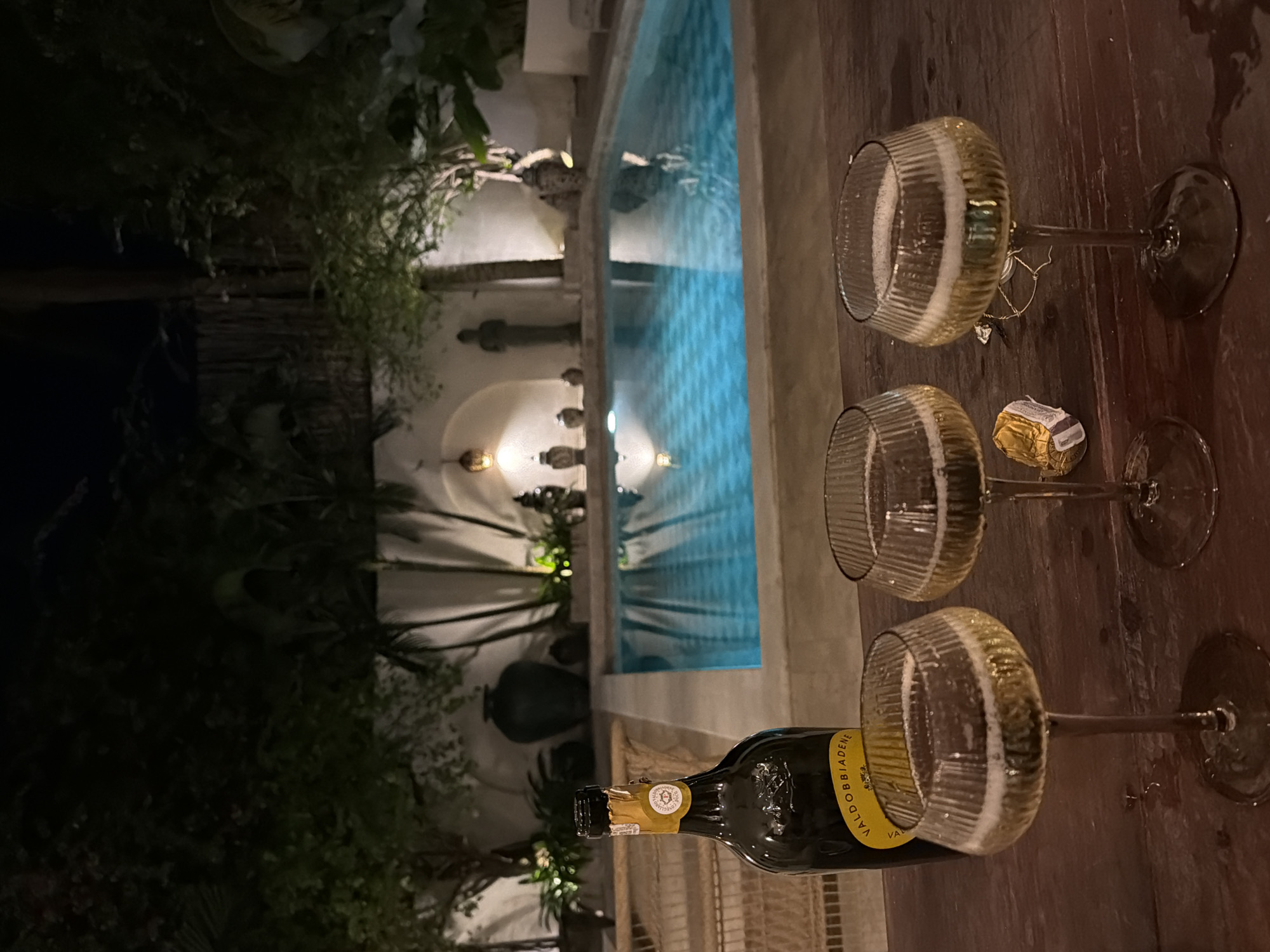 The scene depicts an outdoor setting, likely in the North Kuta region of Indonesia. A swimming pool with turquoise-blue water forms the backdrop, illuminated by subtle lighting emanating from surrounding white architectural elements, possibly columns and structures with decorative statuary. A bottle of Valdobbiadene sparkling wine and three glasses, each partially filled with a bubbly beverage, are arranged on a dark wooden surface. A wrapped candy or truffle is also present. The atmosphere is suggestive of a relaxed, celebratory occasion. Tropical foliage is visible in the background. No people are directly depicted in the frame.