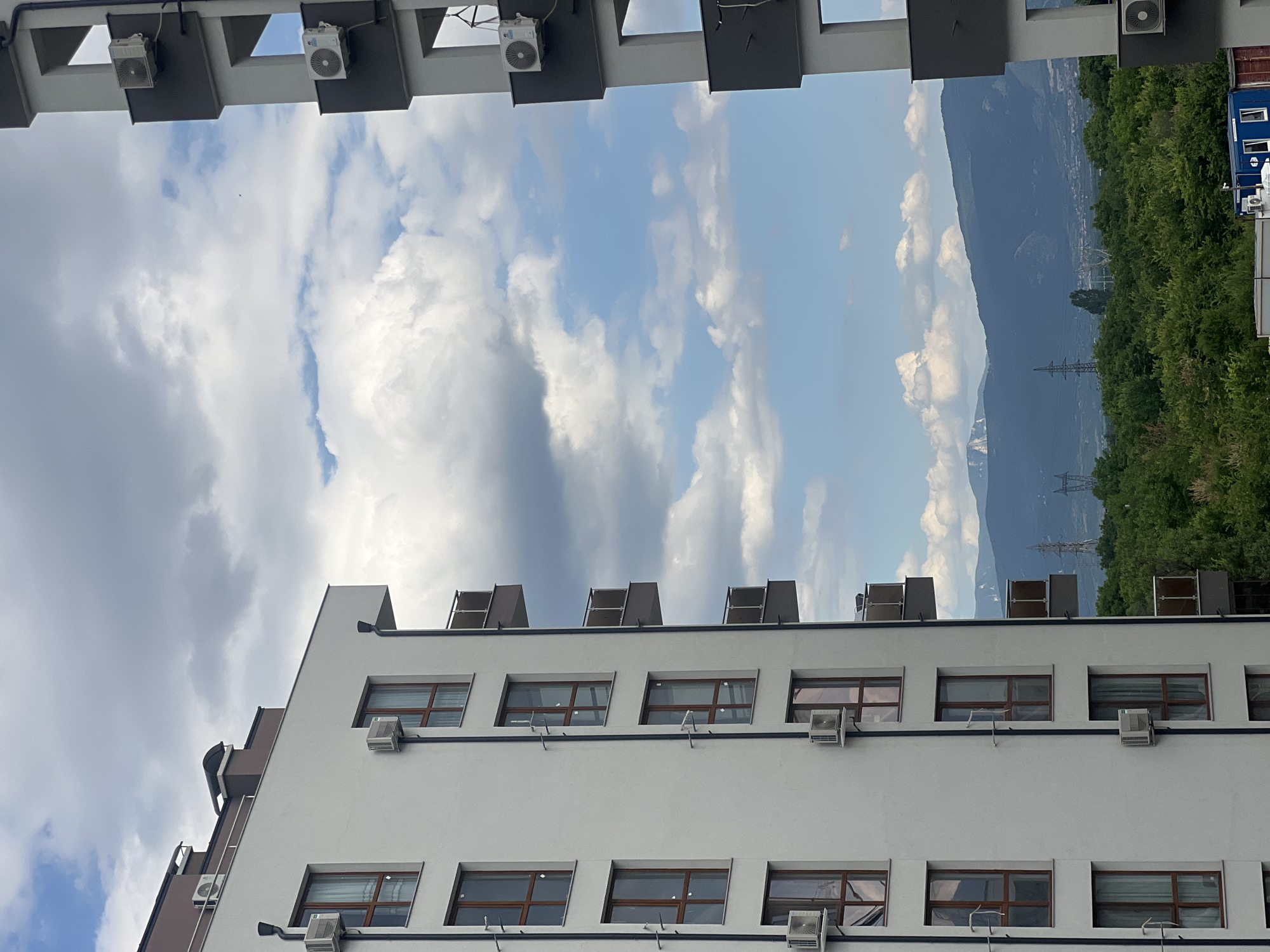 This media file shows a view of a white, modern building with multiple windows and air conditioning units. The building is situated in a landscape with mountains in the background, lush green trees, and a blue sky with scattered clouds. The scene is captured from a slightly low angle, looking up at the building.
The building has a distinctive architectural style, with horizontal lines and large windows that appear to be made of wood and glass. Several outdoor air conditioning units are mounted on the facade of the building, suggesting a temperate climate. On the roof of the building, there are large, rectangular structures that might be ventilation units or part of the building's HVAC system.
In the distance, imposing mountains are visible, some with patches of snow at their peaks. A dense forest borders the mountains, and a few electricity pylons are interspersed within the landscape. The sky is a mix of blue and white clouds, indicating it's daytime, likely late afternoon or early evening, given the lighting. The overall impression is one of a well-maintained urban or suburban environment, possibly a residential or commercial complex, set against a natural backdrop. There is no visible text in the image. The scene evokes a sense of calm and spaciousness, with the natural elements contrasting with the geometric lines of the building.