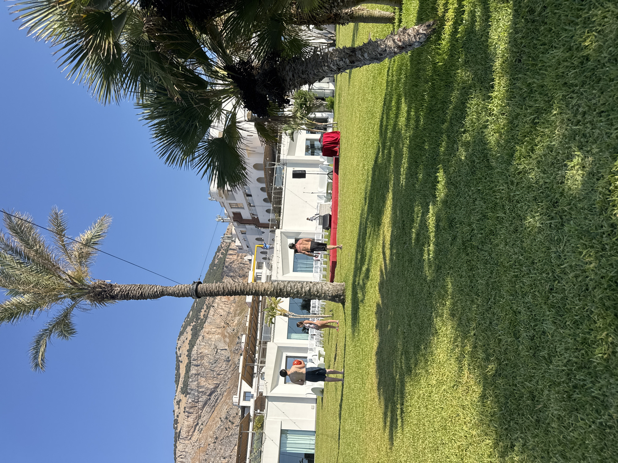 A bright, sunny day is captured at what appears to be a resort or hotel in Isola delle Femmine, Italy. The foreground is dominated by a vibrant green lawn with a subtle slope and dappled shadows from palm trees. In the mid-ground, a group of people are visible in the open air. Two young men, one holding a red ball, stand on the grass. A young woman in a patterned swimsuit is also present. Further back, a stage with red carpet and a red-draped table is set up, with a sound system and chairs nearby. A resort building with white walls and balconies forms the backdrop, and a rocky mountain with sparse vegetation rises behind it. Palm trees are scattered throughout the scene, contributing to the tropical atmosphere. The sky is a clear, vibrant blue.