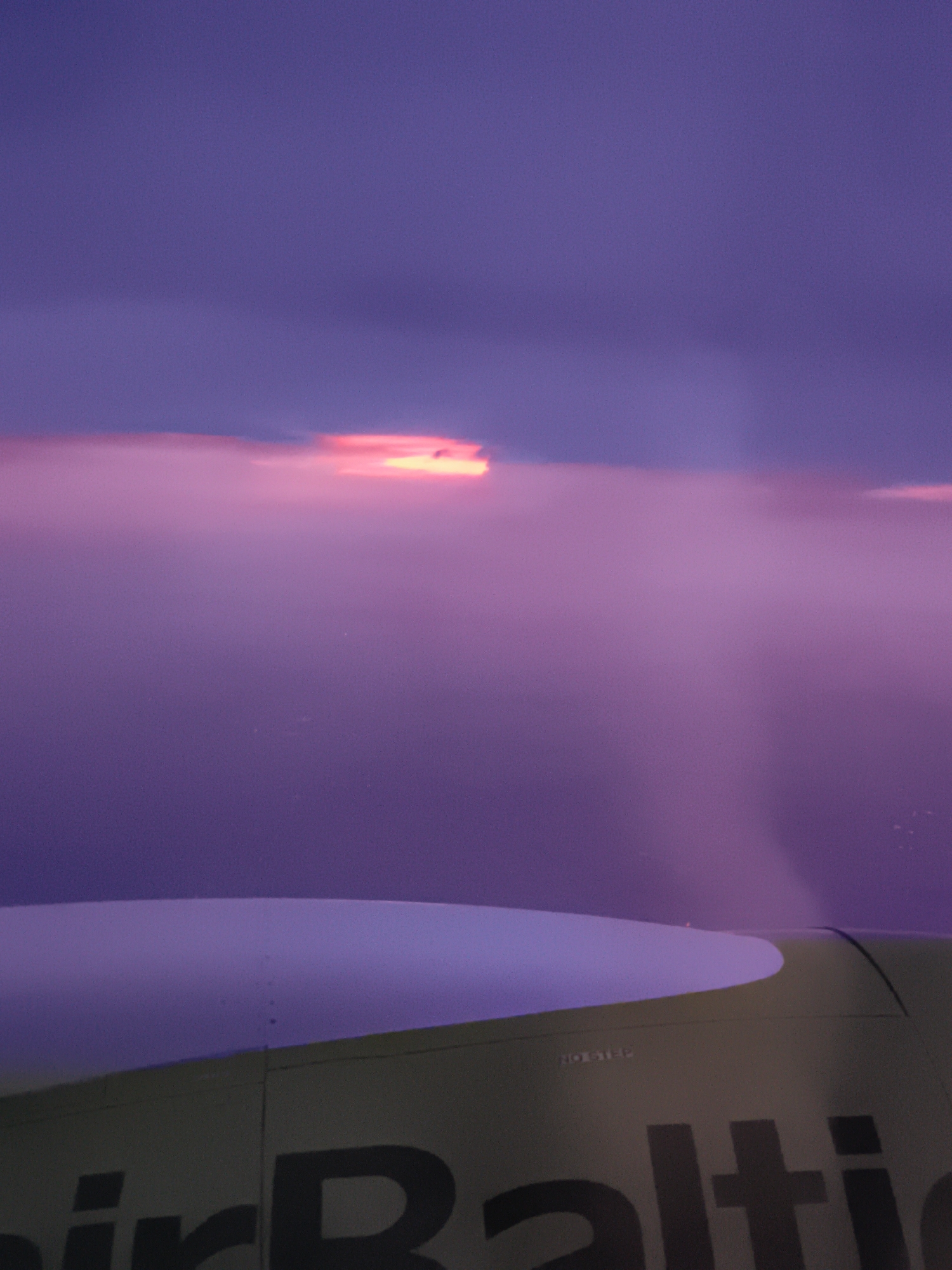 An aerial view captures a sliver of an airplane wing in the foreground, its surface painted a muted olive green with a white curved section. Dark text, likely a logo, is partially visible on the green surface, reading "airBalti". Above the wing, a vast expanse of purple sky dominates the frame, with clouds illuminated by a bright pink and orange sunset or sunrise. The light filters through the clouds, creating a soft, diffused glow. The scene is devoid of people or any identifiable landmarks in Klosterneuburg, Austria.