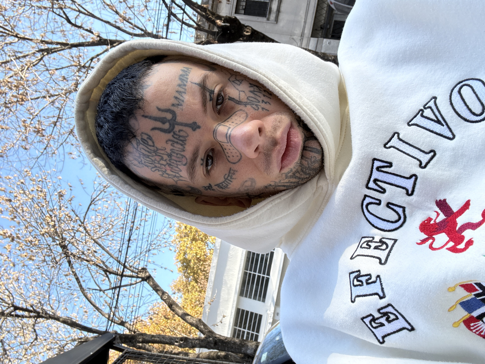 A close-up, tilted selfie of a man with extensive face tattoos wearing a white hoodie with the word "EFECTIVO" vertically embroidered on the front. He has a bandage on his nose. The background includes bare trees against a blue sky, a white building with dark-framed windows, and overhead power lines. Based on the architecture and foliage, the location is likely within the city of Buenos Aires, Argentina.