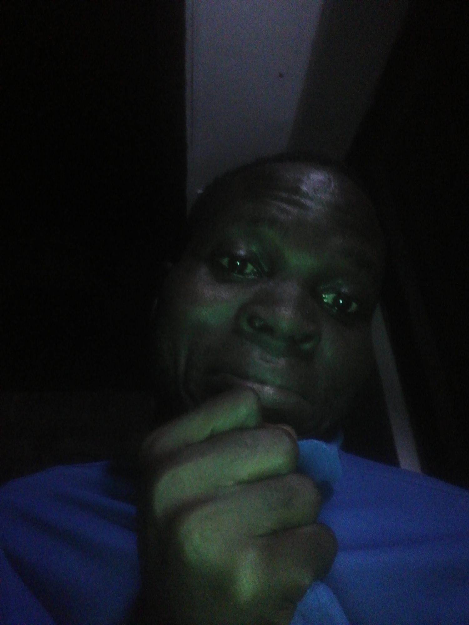 A person with dark skin is centered in the frame with their face directed towards the camera. The person's right hand is positioned near their chin, almost touching it, with a clenched fist. The person is wearing a blue garment. The background is predominantly dark, but there is a lighter, off-white object in the upper-middle to upper-right portion of the background. There are no discernible landmarks or elements in the scene that can definitively identify the location as Ikorodu, Nigeria.