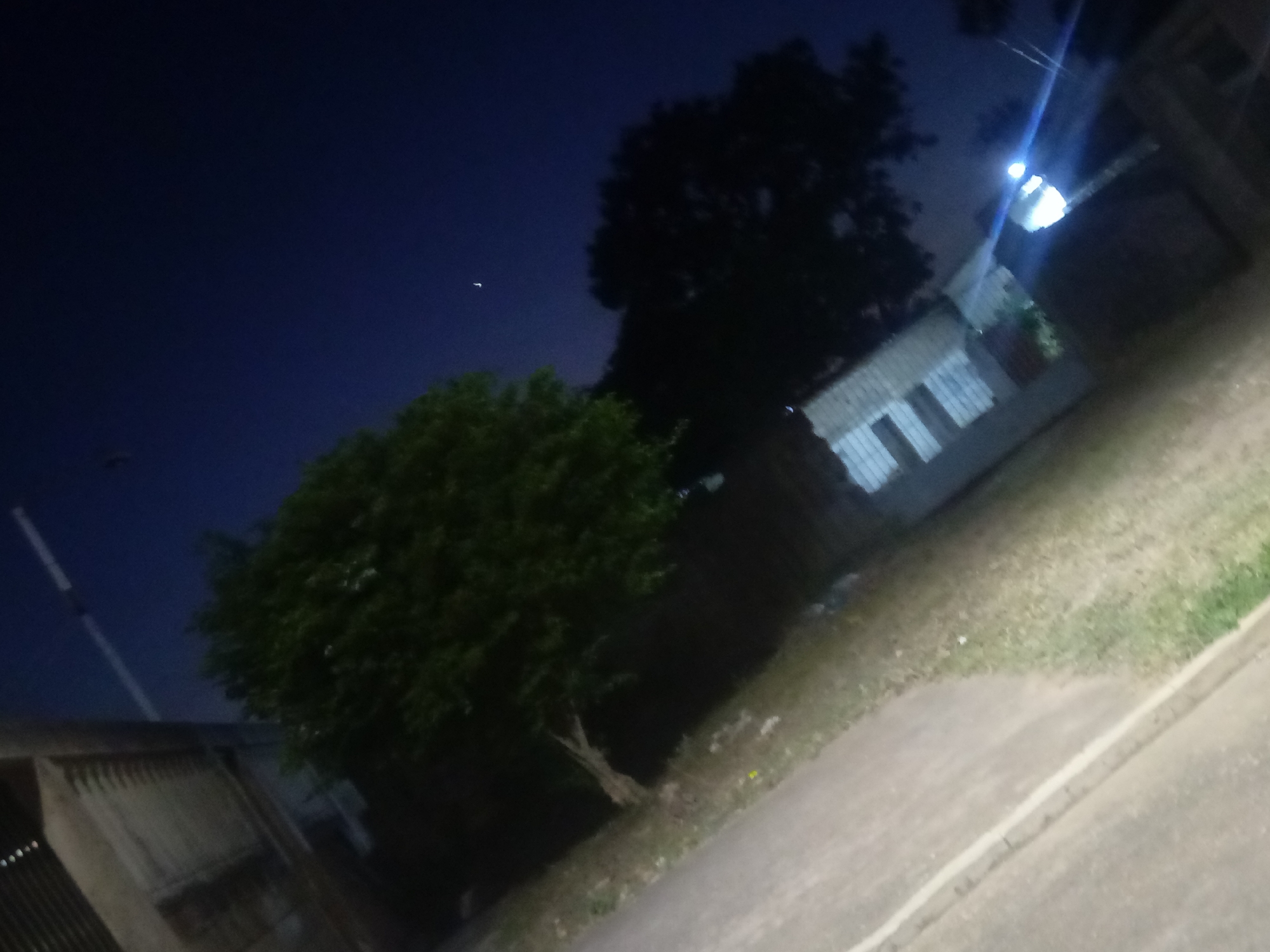 The photograph, taken at night in what is likely Juan Griego, Venezuela, captures a scene dominated by darkness. The sky is a deep blue, punctuated by a single, faint celestial object. Below, a large, dark tree stands to the right of a smaller, green tree. The background features a light-colored building with visible window panes. A street lamp is brightly lit, casting a glow on the surrounding structures and vegetation. A sidewalk runs along the bottom right of the frame, and part of a building with a corrugated metal roof is visible on the bottom left. The overall ambiance suggests a quiet, residential area at night.