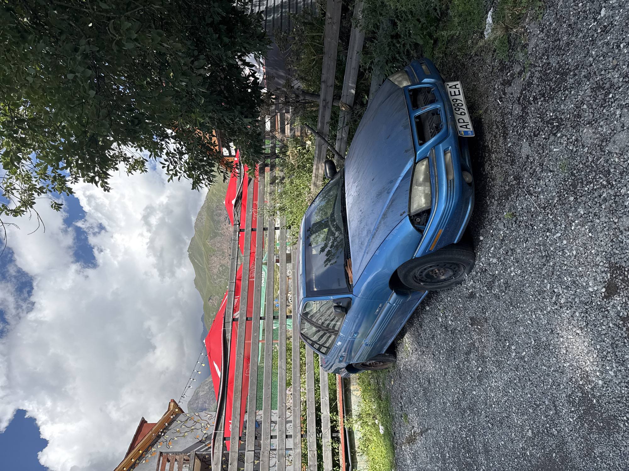An abandoned blue Pontiac Grand Am sedan is parked on a gravel lot in Stepantsminda, Georgia. The car, which appears to be in disrepair with a damaged front end and missing grille, is positioned in front of a wooden fence. Behind the fence, several red umbrellas are visible, suggesting an outdoor seating area for a restaurant or cafe. In the background, lush green mountains under a partly cloudy sky frame the scene. The overall impression is one of rural stillness with a hint of faded functionality.