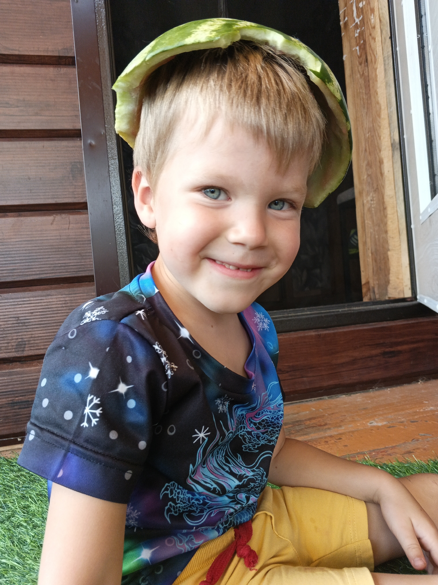 A young Caucasian boy with blond hair and blue eyes sits on green artificial grass outside a wooden structure. He is wearing a black t-shirt with a galaxy and dragon print, and yellow shorts. A piece of watermelon rind is balanced on his head like a hat. The background consists of dark wooden siding, a door frame, and wooden planks on the ground. The image appears to have been taken outdoors in what might be a residential area, possibly in Medvedyovka, Russia, given the architectural style and climate-appropriate clothing.