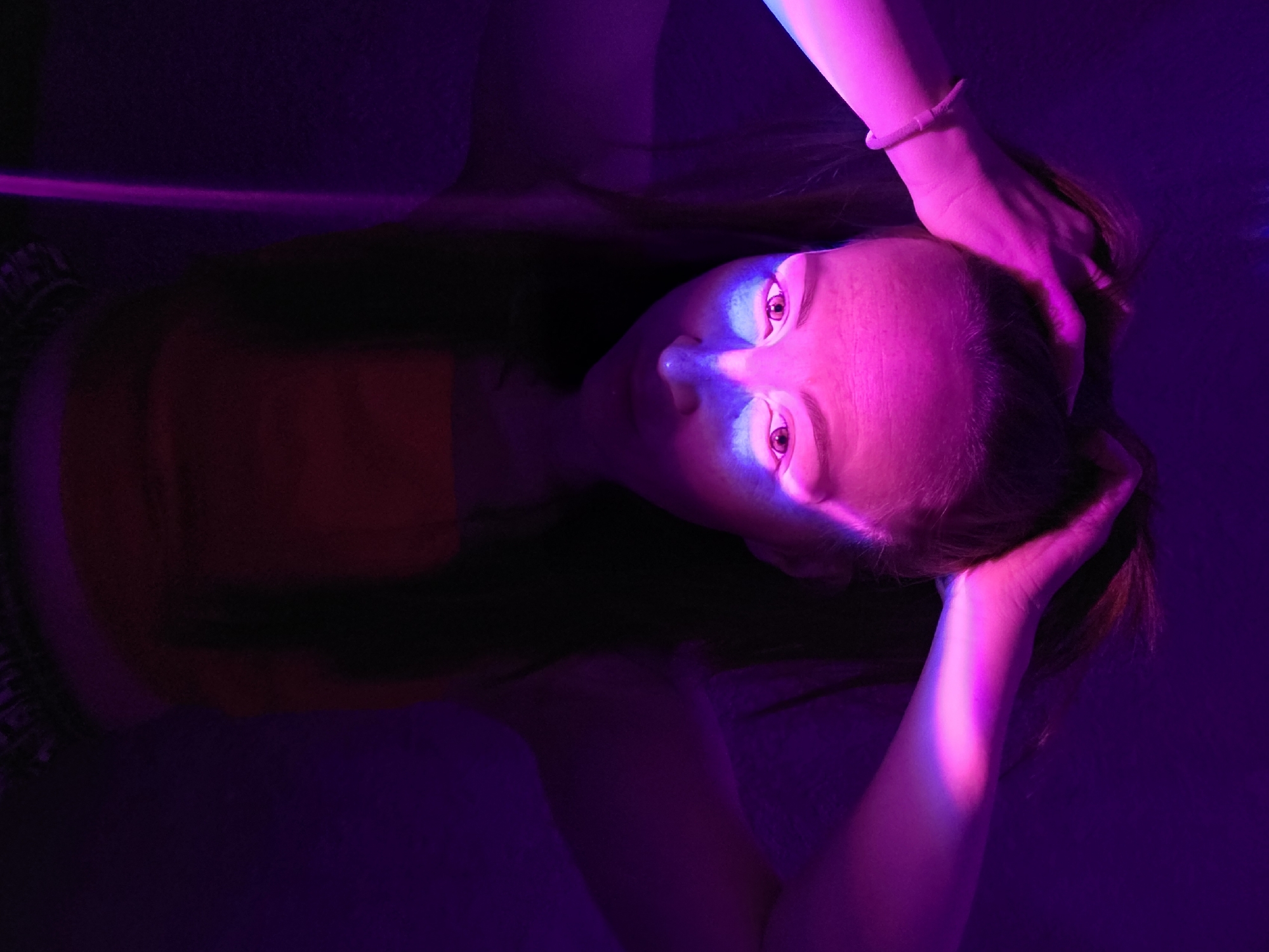 A young woman is depicted from a high-angle, overhead perspective. Her head is tilted back, and she gazes directly at the viewer. Her hands are positioned behind her head, palms touching. She is wearing a short-sleeved shirt and a bracelet on her left wrist. The scene is illuminated with purple and pink lighting, giving her skin a pinkish glow. The background is a dark, saturated purple. No location can be determined from the given picture.