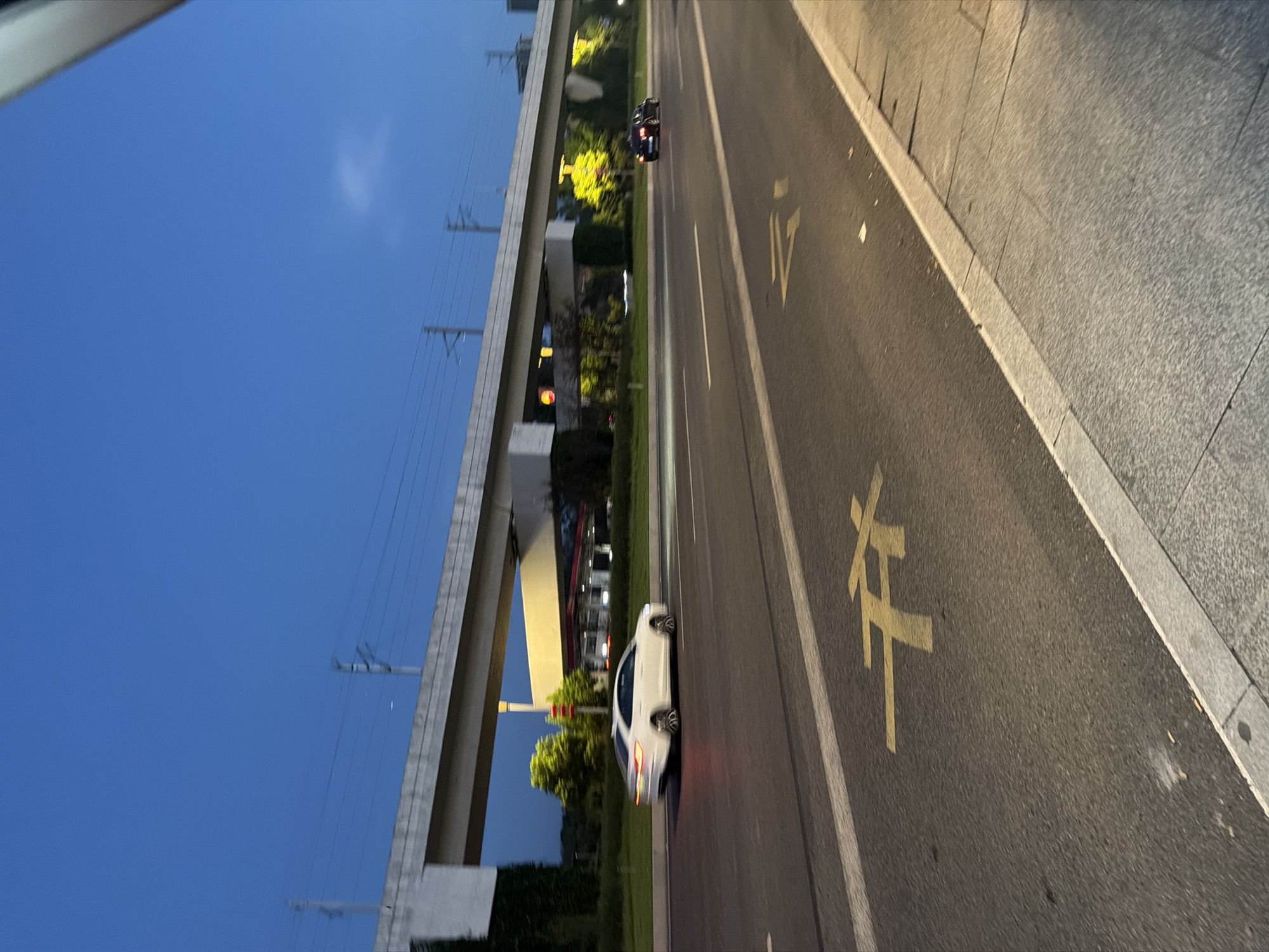 The image depicts a street scene in Jiangjin District, Chongqing, China, at dusk. A white sedan is driving on the right side of the road. In the background, an elevated structure, possibly a bridge or overpass, spans across the roadway. A dark-colored car is also visible further down the road. The sky is a deep blue, indicating the transition from day to night. The road has yellow markings, including what appears to be a symbol or a lane indicator. Trees and buildings are visible beyond the roadway, illuminated by artificial lights. The scene suggests an urban environment with vehicular traffic and infrastructure.