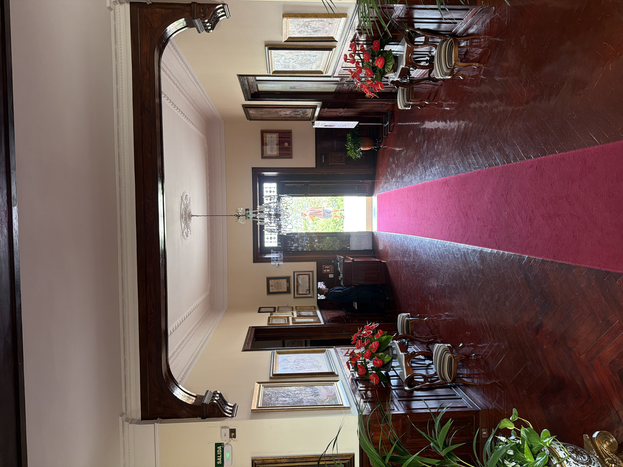 The image shows the entrance hall of a building, likely a historic house or museum, in La Orotava, Spain. A long, red carpet extends down the center of the hall, flanked by a highly polished, dark wood herringbone floor. The walls are a pale yellow and adorned with numerous framed pictures and certificates. On the left, an "EXIT" sign with green lettering is visible. To the right of the entrance, a person in a dark uniform stands behind a reception desk. The hall features ornate wooden arches, decorative ceiling moldings, and a crystal chandelier hanging from the ceiling. Two small chairs with striped upholstery are placed on either side of the hall, near decorative tables with floral arrangements. In the background, double doors open to an outdoor area with greenery.