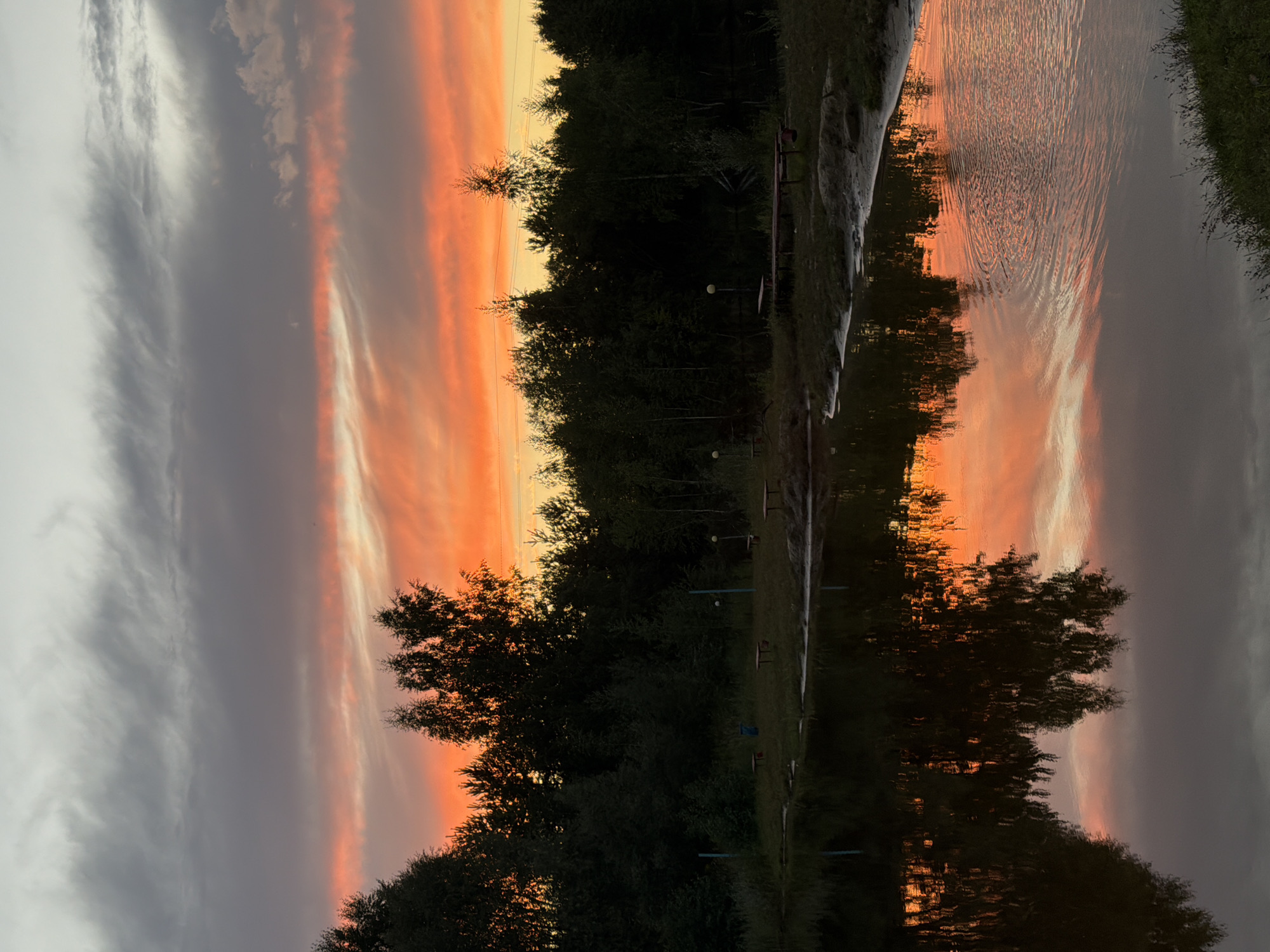 The image captures a sunset over a body of water, likely a lake or pond, bordered by a dense line of trees. The sky is painted with dramatic colors, transitioning from a pale gray at the top to vibrant oranges and pinks closer to the horizon. Wispy clouds stretch across the sky, reflecting the sunset hues. In the foreground, the water's surface is calm, mirroring the colorful sky and the dark silhouettes of the trees. Along the bank, there are signs of human activity: several wooden benches and tables are visible, suggesting a park or recreational area. A blue trash can is also present. The overall impression is one of a serene, natural landscape enhanced by the beauty of a sunset, located in the vicinity of Глубоково, Russia.