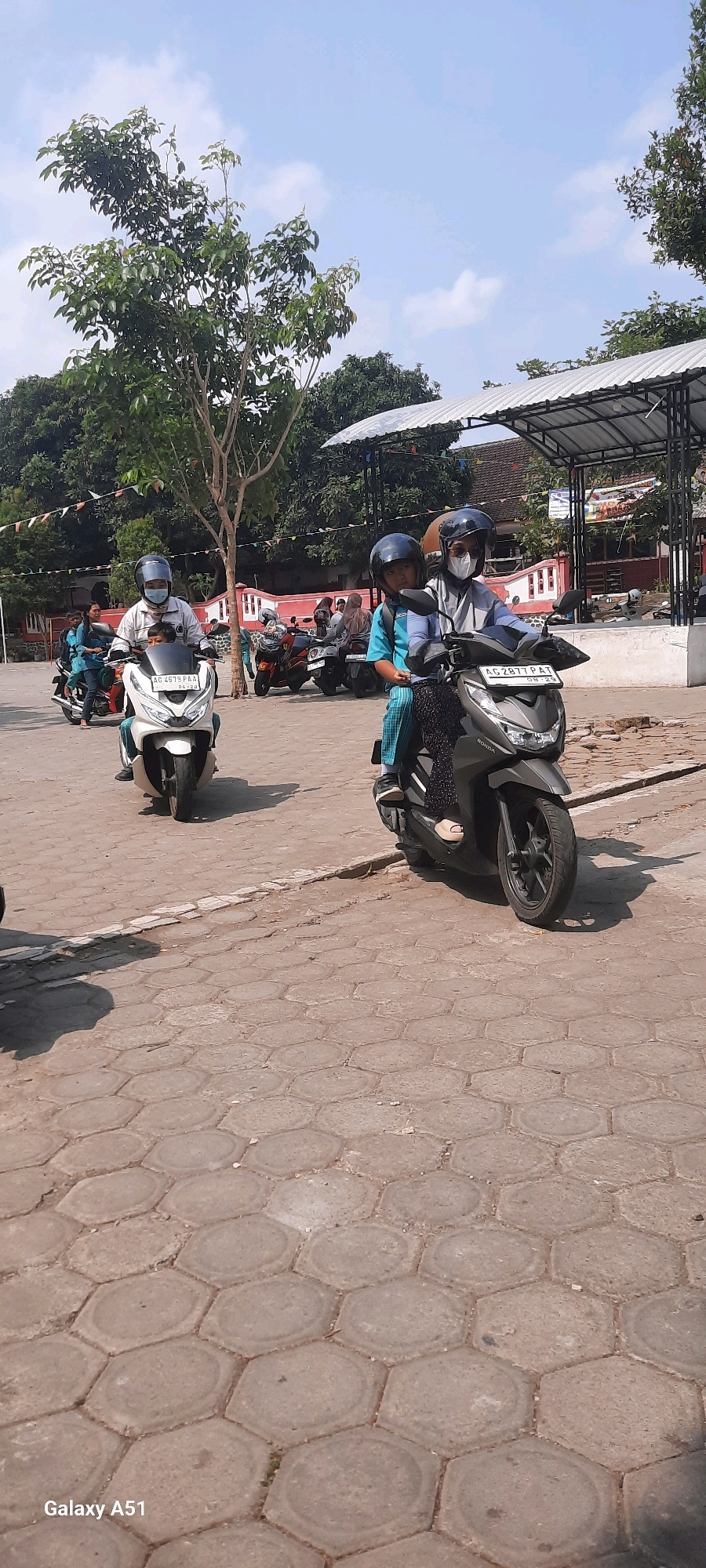 In Tangkil, Indonesia, under a bright, partly cloudy sky, two motorcycles with riders are captured in motion on a paved area. The motorcycle in the foreground is dark gray, with a rider wearing a helmet and a light gray face mask, along with a passenger in a blue shirt and a helmet. They are moving from left to right. Behind them, a white motorcycle is also in motion, with a rider in a helmet and a light-colored jacket, carrying a passenger in a blue top. Other motorcycles and people are visible in the background, suggesting a public space or thoroughfare. Trees line the background, and a covered structure with a white, corrugated roof is present on the right side of the frame. The ground is paved with hexagonal-shaped interlocking blocks.