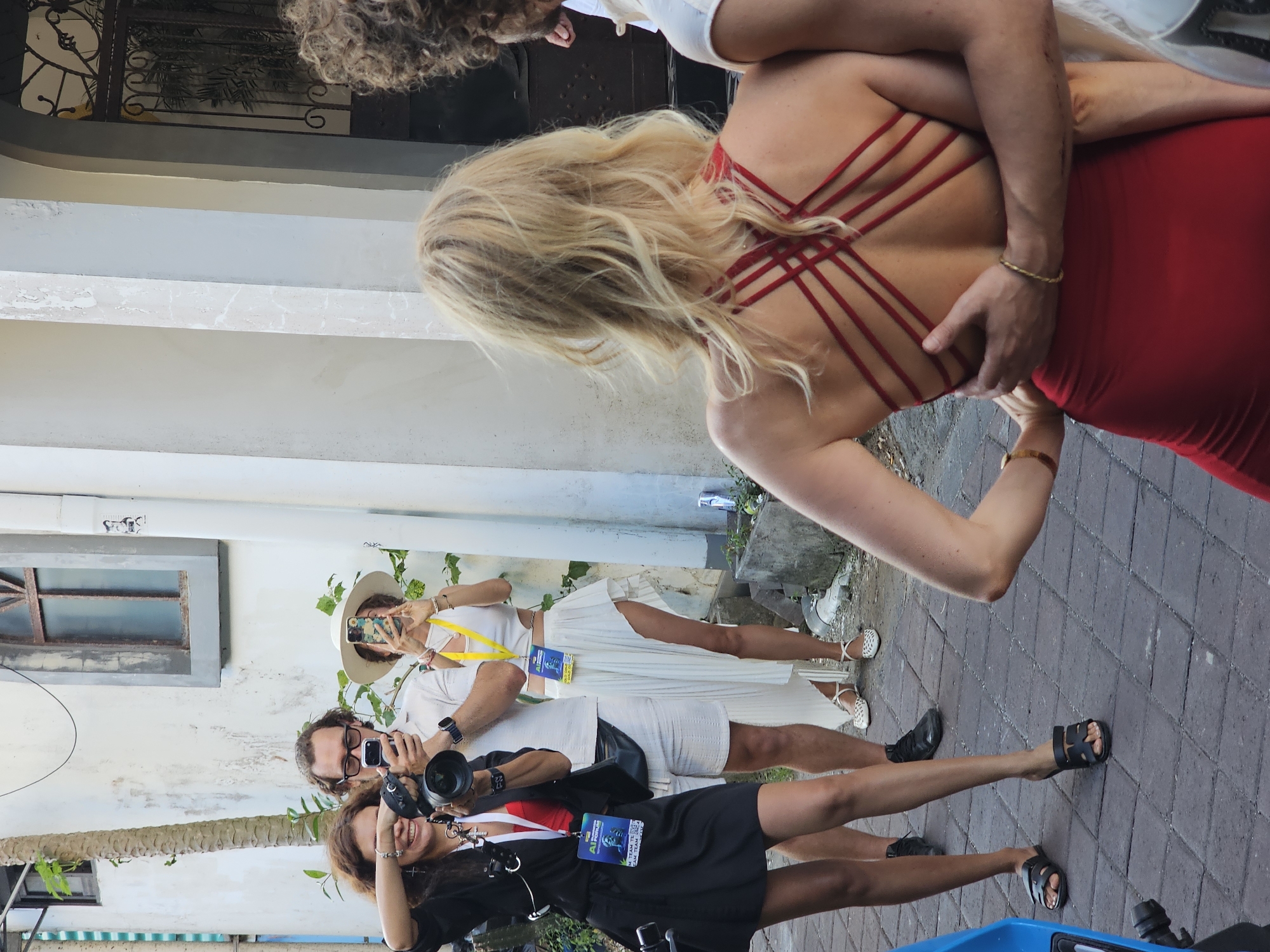 A group of people are on a stone paved road in North Kuta, Indonesia. A blonde woman is being hugged by a man. She is wearing a red dress. Three people are standing near her. A woman is taking a photo with a cell phone, and a man and another woman are holding cameras. All three are wearing lanyards with identification badges. The road is next to a light gray building with a window. Some foliage is present near the building.