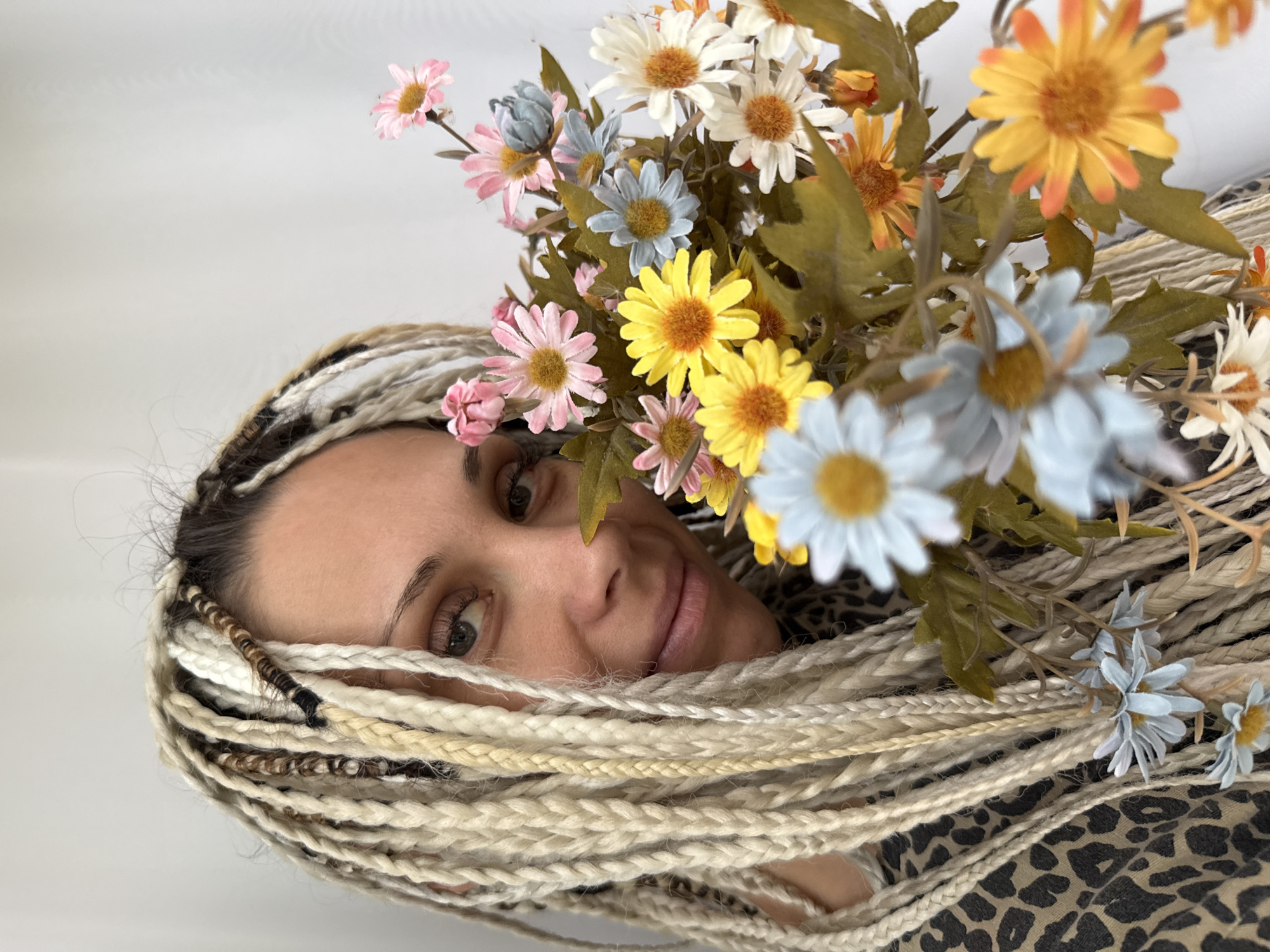 A close-up, medium shot shows a woman with blonde-toned braided hair, accessorized with small blue and white flowers, holding a bouquet of artificial daisies. She is positioned on the left side of the frame, her face turned towards the viewer, with her right eye in focus and her left eye partially obscured. The bouquet, a mix of yellow, pink, light blue, white, and orange daisies, is held in the foreground, to the right of the woman. The background is a plain, light-colored, textured surface, possibly a wall or fabric. The woman's attire is a leopard-print top. The image was captured in the city of Rusanovo, Russia.