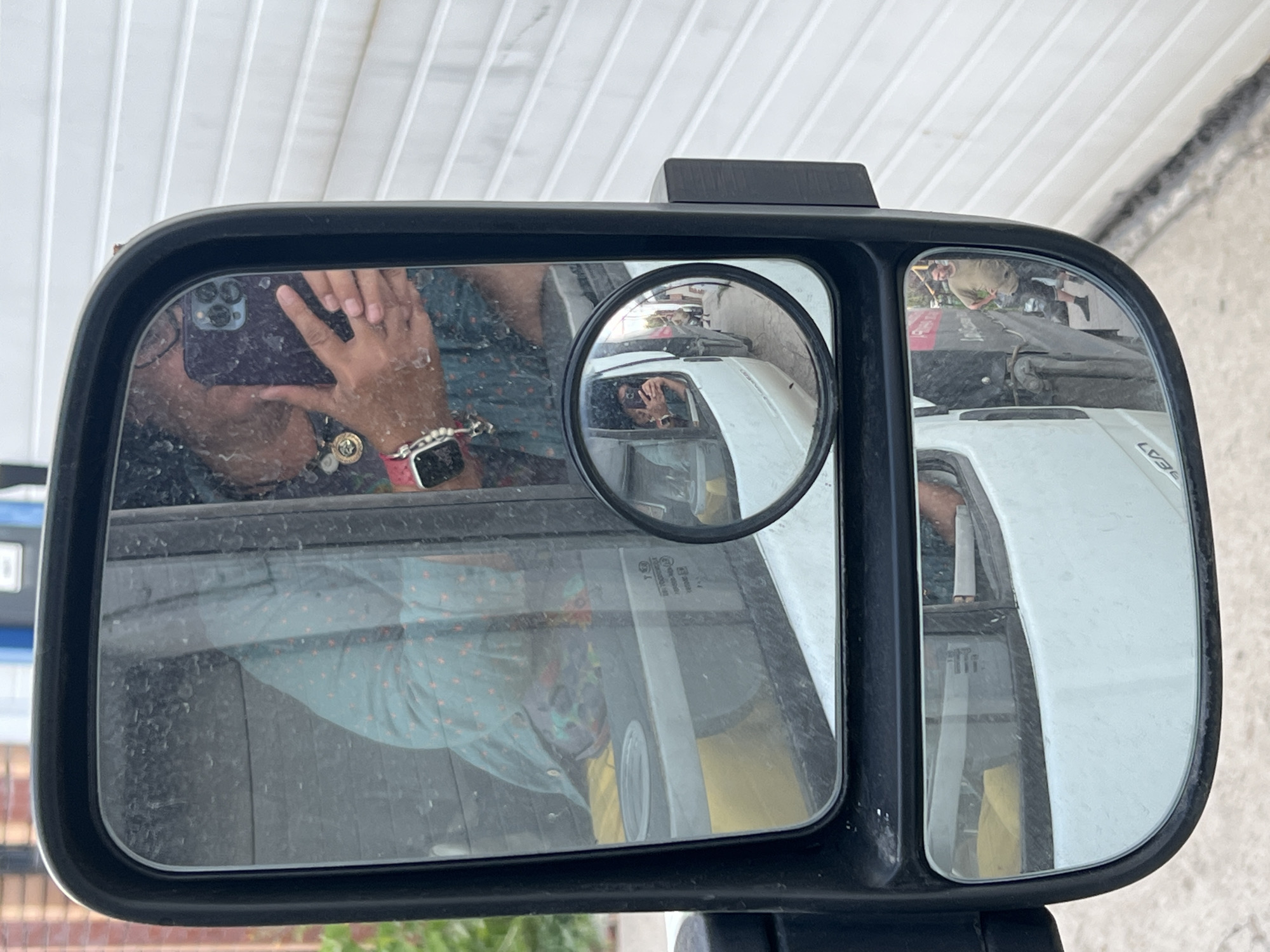 The image shows a reflection in a side-view mirror of a vehicle. In the main mirror, a person is visible, taking a selfie with a smartphone. They are wearing a blue shirt with a pattern and a watch. There is also a reflection of the interior of the vehicle, including a yellow object and a cup holder.
A smaller circular mirror, attached to the main mirror, shows a further reflection. This reflection depicts the driver of another vehicle, also taking a photo with a smartphone, and the road stretching ahead with other vehicles and buildings on the side.
The background outside the mirror is a white, textured wall, possibly a building's facade.
The location is identified as Ozernyy, Russia. The scene appears to be an urban or semi-urban setting, possibly a parking lot or a street. The overall atmosphere is neutral, focusing on the act of taking reflections.