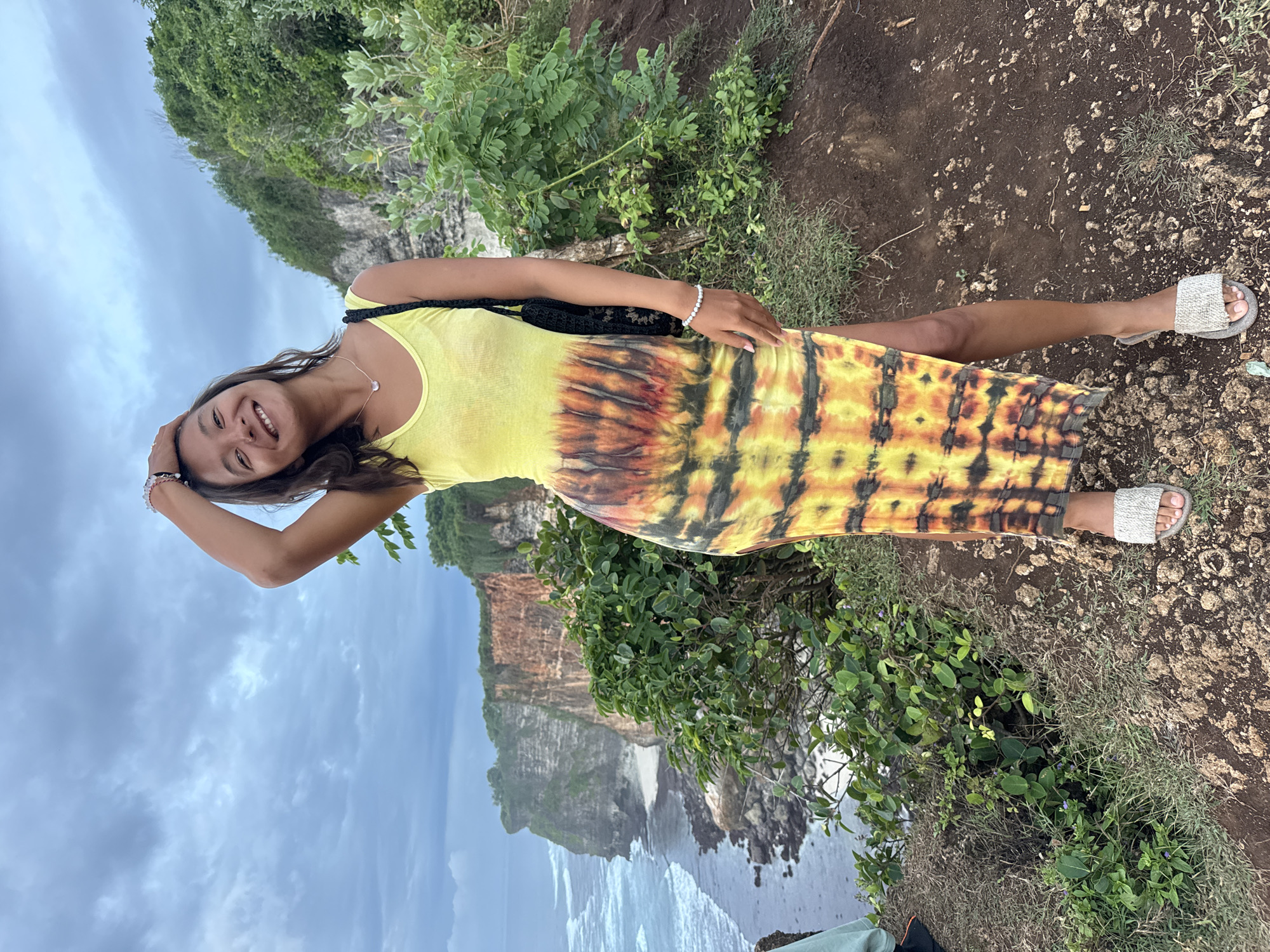 A woman in a yellow and orange tie-dye dress stands on a rocky cliff overlooking the ocean in Kuta Selatan, Indonesia. She has long dark hair and is smiling while holding her hand on her head. She wears a necklace and bracelets on both wrists. Her sandals are white. Behind her is a rocky coastline with lush green vegetation. The sky is overcast.