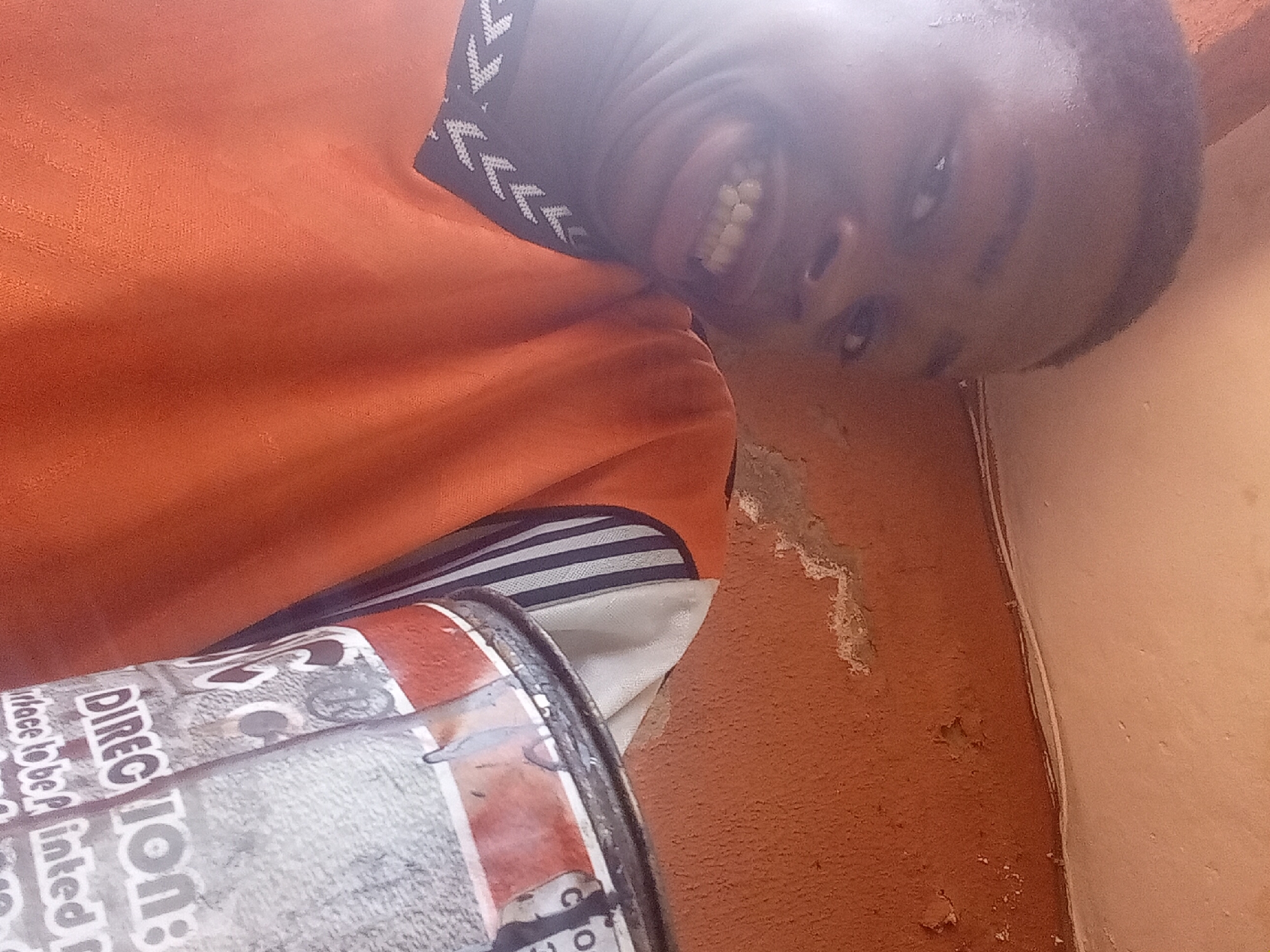 A close-up indoor shot features a person smiling with their teeth showing. The person is wearing an orange shirt with a patterned collar. To the side of the person, a can with writing on it is visible. The background includes a textured orange wall and what appears to be a corner where two walls meet. The environment suggests an interior space, likely in Nkpor, Nigeria, based on the provided context. The focus is on the person's face and smile, with the can and the background providing additional context to the scene.