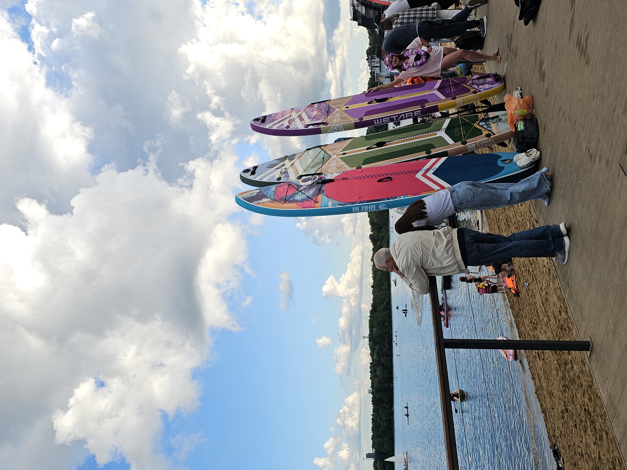 The image captures a view of the Khimki Reservoir in Khimki, Russia, on a cloudy day. In the foreground, a wooden boardwalk runs along the edge of a sandy beach. Several people are present, some standing and observing the water, while others are preparing for or engaged in water activities. Three colorful stand-up paddleboards are propped upright on the boardwalk.
An elderly man with gray hair is leaning on a railing, looking out at the reservoir. To his left, a young woman in jeans and a white t-shirt stands near one of the paddleboards. To the right of the paddleboards, a woman wearing a pink lei and a pink t-shirt is posing for the camera. Other individuals are visible in the background, including some on paddleboards in the water and people walking on the beach.
The reservoir itself is calm, with several small boats and paddleboards dotting its surface. The opposite shore is lined with a dense green forest, and buildings and industrial structures can be seen in the distance. The sky is a mix of blue and white clouds, suggesting a partly cloudy day.