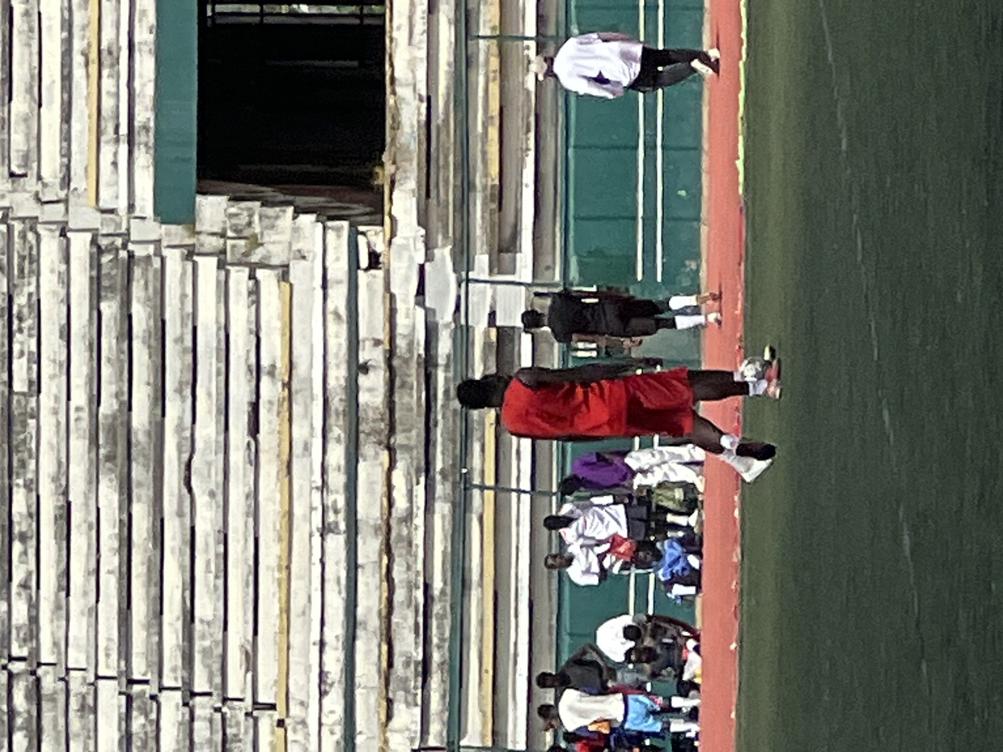 This image captures a scene at the New Stadium in Nigeria, likely during a football (soccer) training or practice session. In the foreground, a person wearing a red sleeveless top and red shorts is walking across a green field. Another person in a black shirt and shorts is walking away from the viewer. To the right, a person in a white shirt and black pants is also walking. In the middle ground, several individuals are visible in the background, some standing and others sitting on the ground. They are wearing various athletic attire. Behind them, a grandstand with rows of weathered seats stretches across the frame, indicating the stadium's setting. The overall atmosphere suggests a sporting event or training in progress.