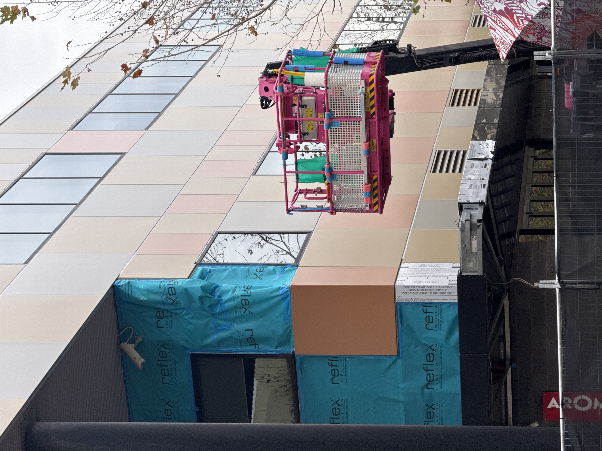 In Perth, Australia, a construction site features a multi-story building with a facade of light-colored vertical panels in shades of cream, beige, and pale pink, interspersed with narrow, horizontal windows. A pink aerial work platform is positioned in front of the building, its basket containing various tools and materials, including green fabric sheets, yellow poles, and a roll of white paper. One section of the building's exterior is covered in a bright turquoise wrap, with "reflex INSULATION" printed on it in black text. A security camera is visible mounted on the underside of an overhang. Below the building, a black metal fence encloses the construction area. A red and white patterned tarp is visible on the right side of the frame, and a red sign with white lettering reading "AROM" is attached to a fence post on the left. The sky is overcast.