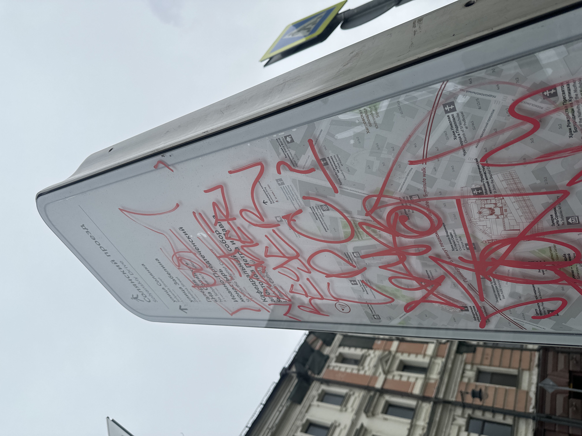 The image captures an upward, tilted view of a public informational sign, likely a map, against an overcast sky. The sign's clear protective covering is heavily defaced with bright red, chaotic graffiti, making much of the underlying content difficult to read. The scribbles appear random rather than forming legible words.
Beneath the prominent red markings, a detailed urban map is visible, featuring street layouts and points of interest with text in both Russian and English. Readable place names include "Солянский проезд" (Solyanskiy Drive), "улица Солянка" (Solyanka Street), "улица Забелина" (Zabelina Street), and "Хитровская площадь" (Khitrovskaya Square). Other landmarks visible on the map include "Высшая школа экономики (НИУ ВШЭ)" (Higher School of Economics), "Иоанно-Предтеченский женский монастырь" (St John the Baptist Convent), "Кафедральный собор апостолов Петра и Павл" (Lutheran Cathedral of Saints Peter and Paul), and several churches like "Храм Николая Чудотворца в Подкопаях" (Church of St Nicholas at Podkopai). Directions like "Примерно 5 минут пешком" (Approx. 5-minute walk) are also present.
In the upper background, a faded yellow pedestrian crossing sign is partially visible. Below the informational sign, a section of an old building with multiple rectangular windows and a light-colored facade is visible, suggesting an urban setting. The grey, cloudy sky indicates an overcast day. The prominent graffiti signifies an act of vandalism on public property.