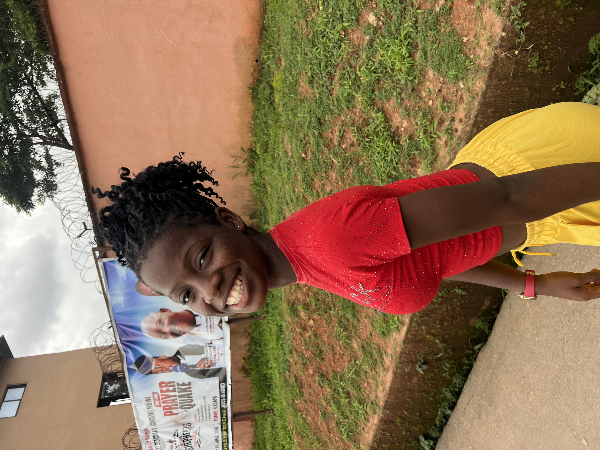 A young Black female with dark skin and dark braided hair is smiling at the camera, looking to her left. She is wearing a short-sleeved red crop top adorned with small rhinestones and bright yellow drawstring pants. A pink watch is visible on her left wrist. The background features a beige building with two windows, a large banner with text and images of people, and a light orange wall topped with barbed wire. Lush green vegetation and a pathway are also present. The banner advertises a "PRAYER QUAKE" gathering in Nnewi, Nigeria, with specific dates and times mentioned.