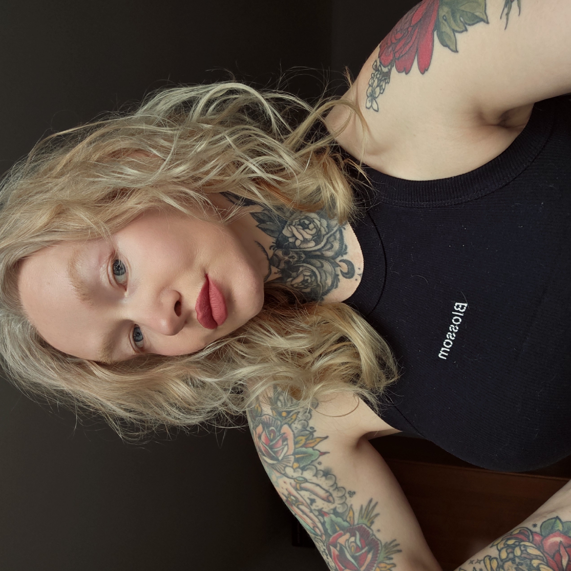 A woman is angled in the frame, facing the camera. She has fair skin, blonde curly hair, and blue eyes. She wears a black tank top with the word "Blossom" printed vertically on it. Multiple tattoos adorn her arms and neck. A dark wall is visible in the background, suggesting an indoor setting. The location in the city of Saint Petersburg, Russia is undetermined.