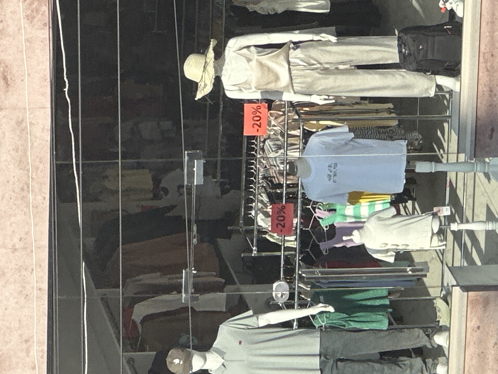 The image depicts a storefront display of clothing on mannequins in Gyumri, Armenia. On the left, a male mannequin wears a grey polo shirt, a brown baseball cap, and grey pants. To its right, a female mannequin is dressed in a beige satin tank top and matching wide-leg pants, topped with a straw hat. In the background, racks of clothing are visible, some with "-20%" sale signs. Another smaller mannequin in the foreground displays a white t-shirt. A black backpack rests on the floor near the female mannequin. The storefront appears to be a clothing boutique or department store.