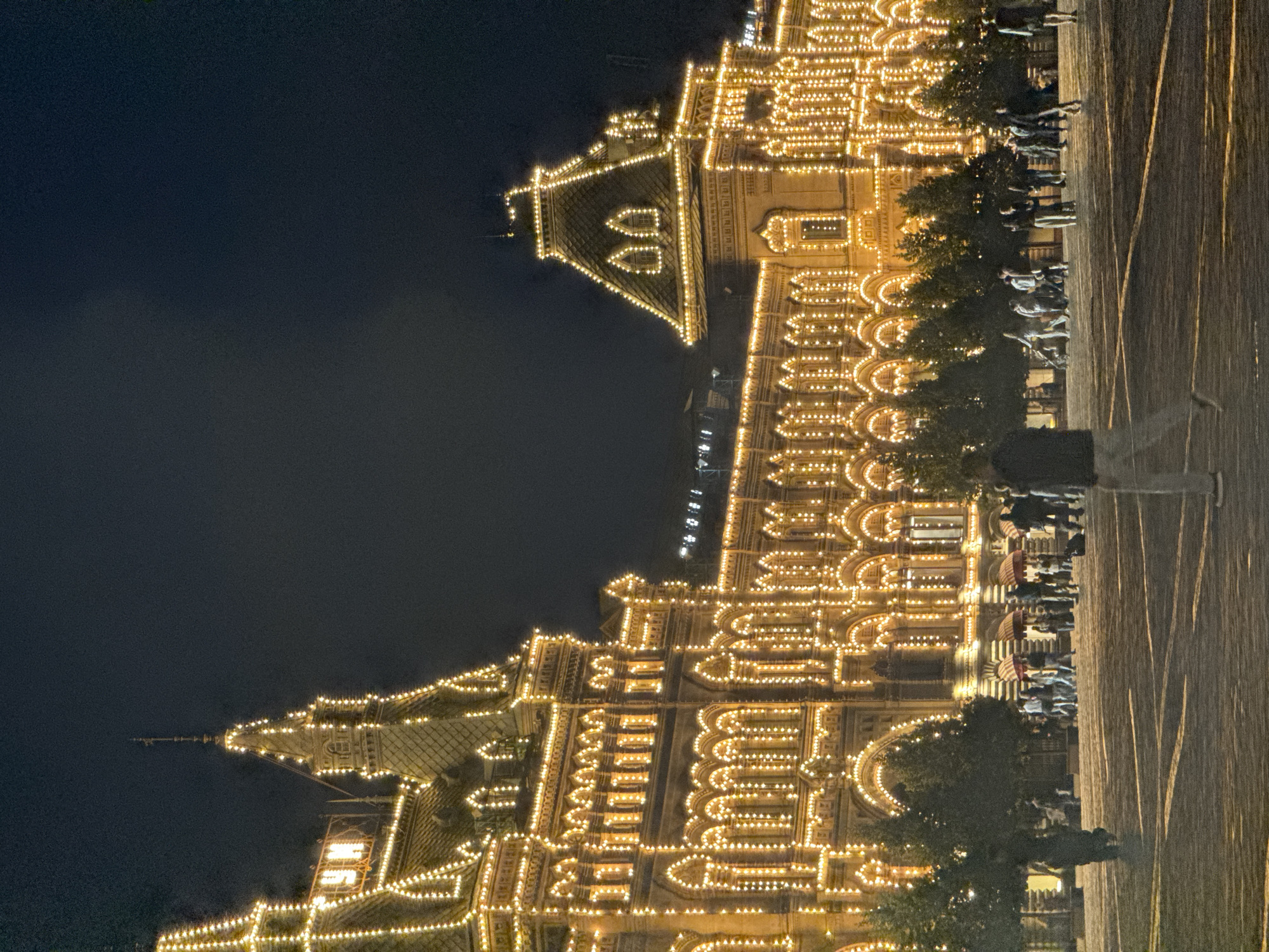 The image depicts GUM department store on Red Square in Moscow, Russia, at night. The building is illuminated with numerous strings of warm white lights that outline its architectural features, including arched windows, cornices, and turrets. The sky above is dark, indicating nighttime. In the foreground, a cobblestone plaza is visible, with several people walking and standing. The people are diverse in age and attire, with some walking in groups and others individually. Shadows from the building and people are cast across the plaza. Trees line the area in front of the building. The overall scene conveys a festive and illuminated urban environment at night.