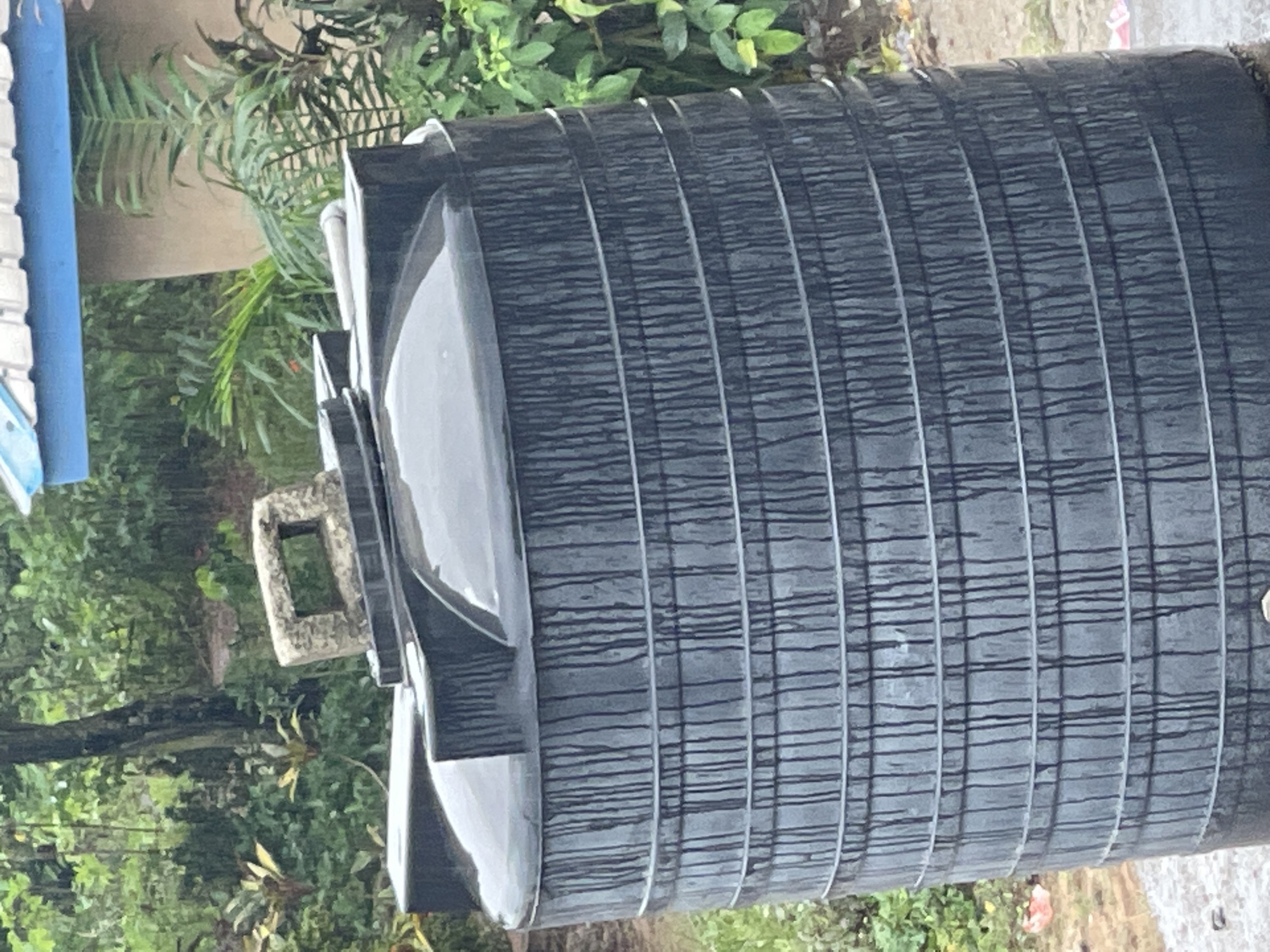 A large, black, cylindrical plastic water storage tank sits outdoors in what appears to be a lush, green, overgrown area, likely within a residential or semi-rural setting in Abraka, Nigeria. The tank has numerous circumferential rings or bands, possibly for structural reinforcement, and is wet, with water droplets or streaks visible on its surface. It has a raised section on its left side, with what appears to be a valve or connection point. The surrounding environment includes various plants and foliage, suggesting a tropical or subtropical climate. A portion of a building with a blue roof is visible in the background. The scene suggests a rainy or recently rainy condition.