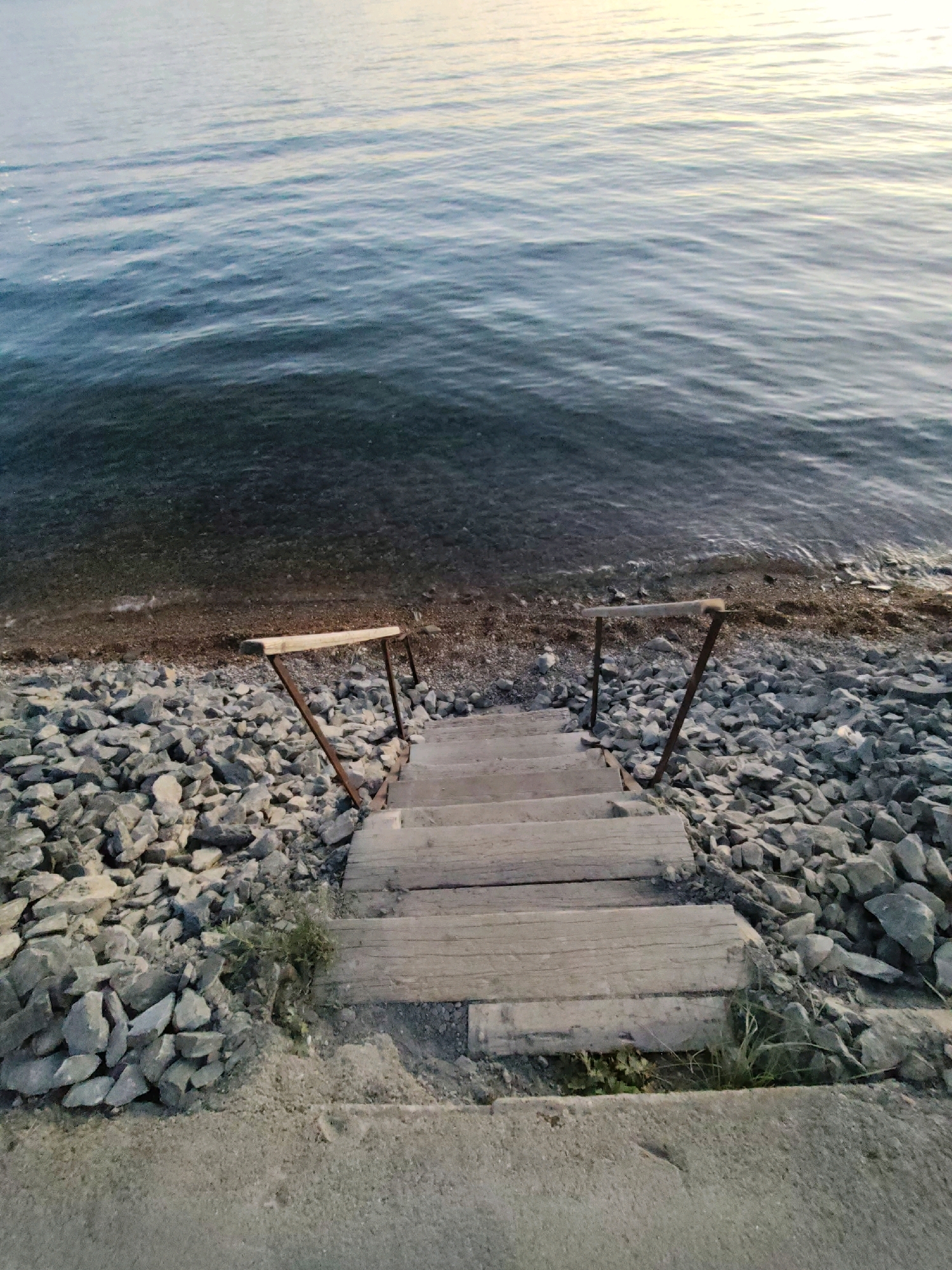 An aerial perspective captures a rustic wooden staircase descending towards the calm, rippling waters of a lake. The stairs are constructed of weathered planks and flanked by a simple wooden handrail supported by rusted metal posts. The base of the staircase is surrounded by a shoreline of grey, jagged rocks and pebbles. Beyond the rocks, the dark, tranquil water extends to the horizon, reflecting a soft, pale yellow light from what appears to be a setting sun. The environment suggests a natural, unmanicured waterfront, likely a remote or less developed area.