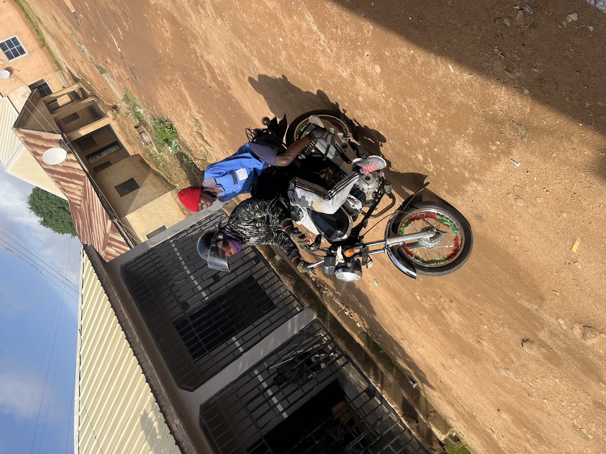 Two individuals are riding a motorcycle on a dirt road in Gura Top, Nigeria. The person in front is wearing a black jacket with a patterned design, gloves, and a clear-visored helmet. The person behind is wearing a blue long-sleeved shirt, dark shorts, and a red beanie. The motorcycle is a silver and black model with decorative green and red accents on the front wheel. The environment features buildings with corrugated metal roofs and satellite dishes, indicative of a residential area. The road is unpaved and dusty, with scattered debris. The sky is visible with some clouds.